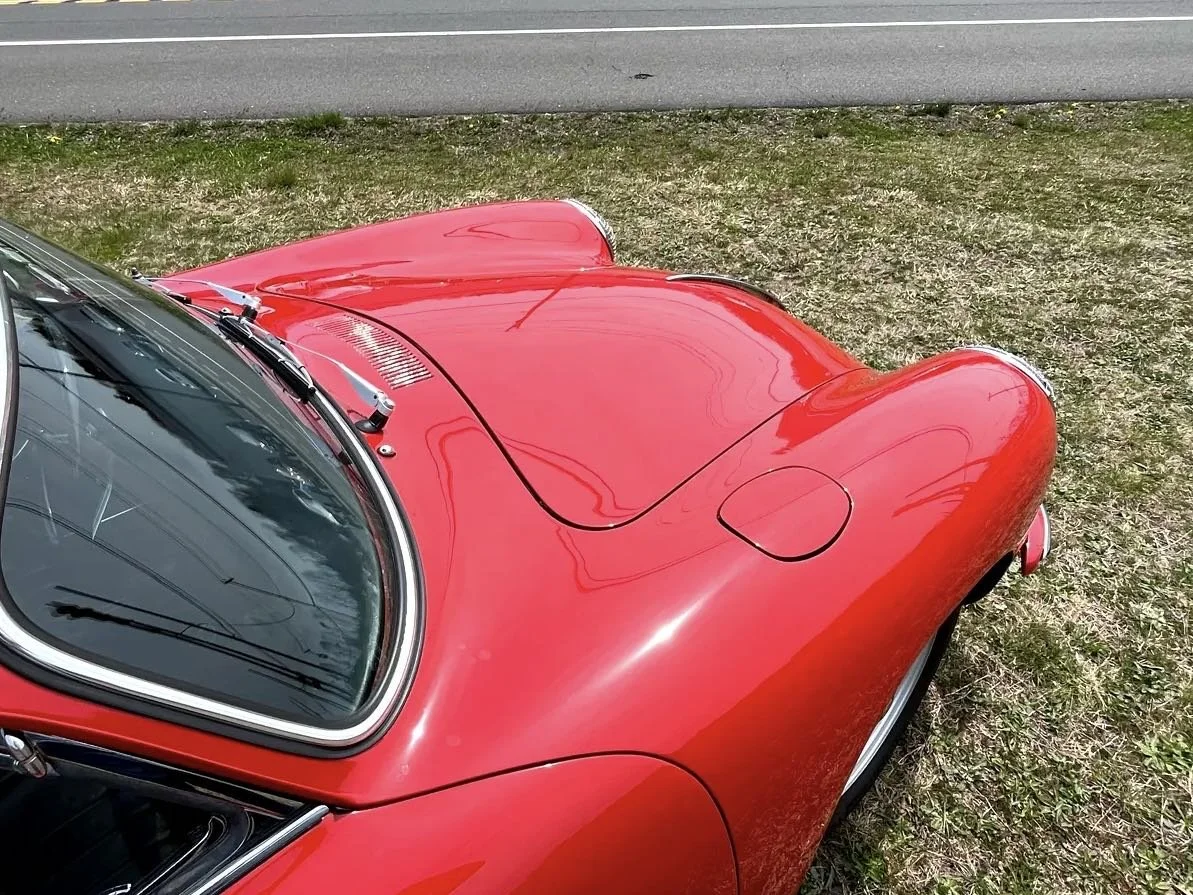 Close-up view of a vintage red sports car parked on grass near a road, showing the windshield, hood, and part of the front wheel.