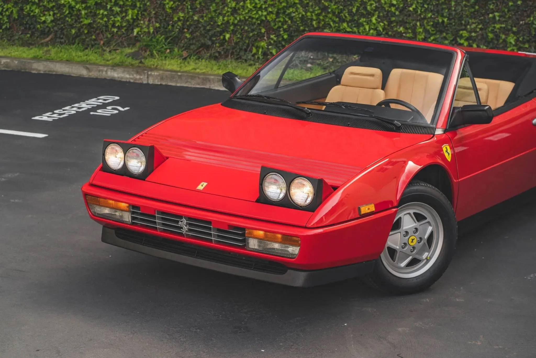 Red vintage Ferrari convertible with pop-up headlights parked in a lot near a hedge, beige interior visible.