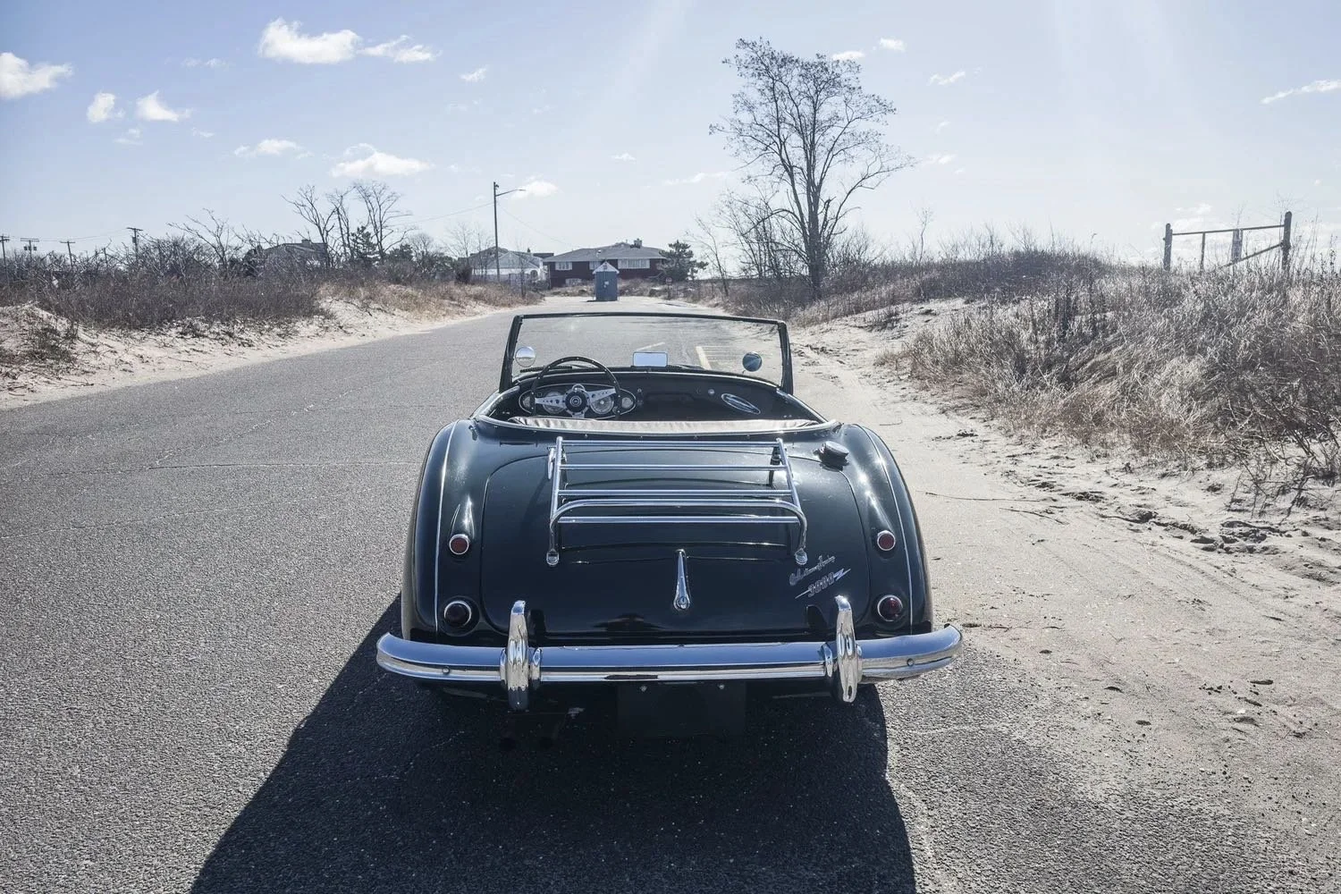 A vintage black convertible car parked on a sandy road near a beach, with a house, trees, and a blue sky in the background.