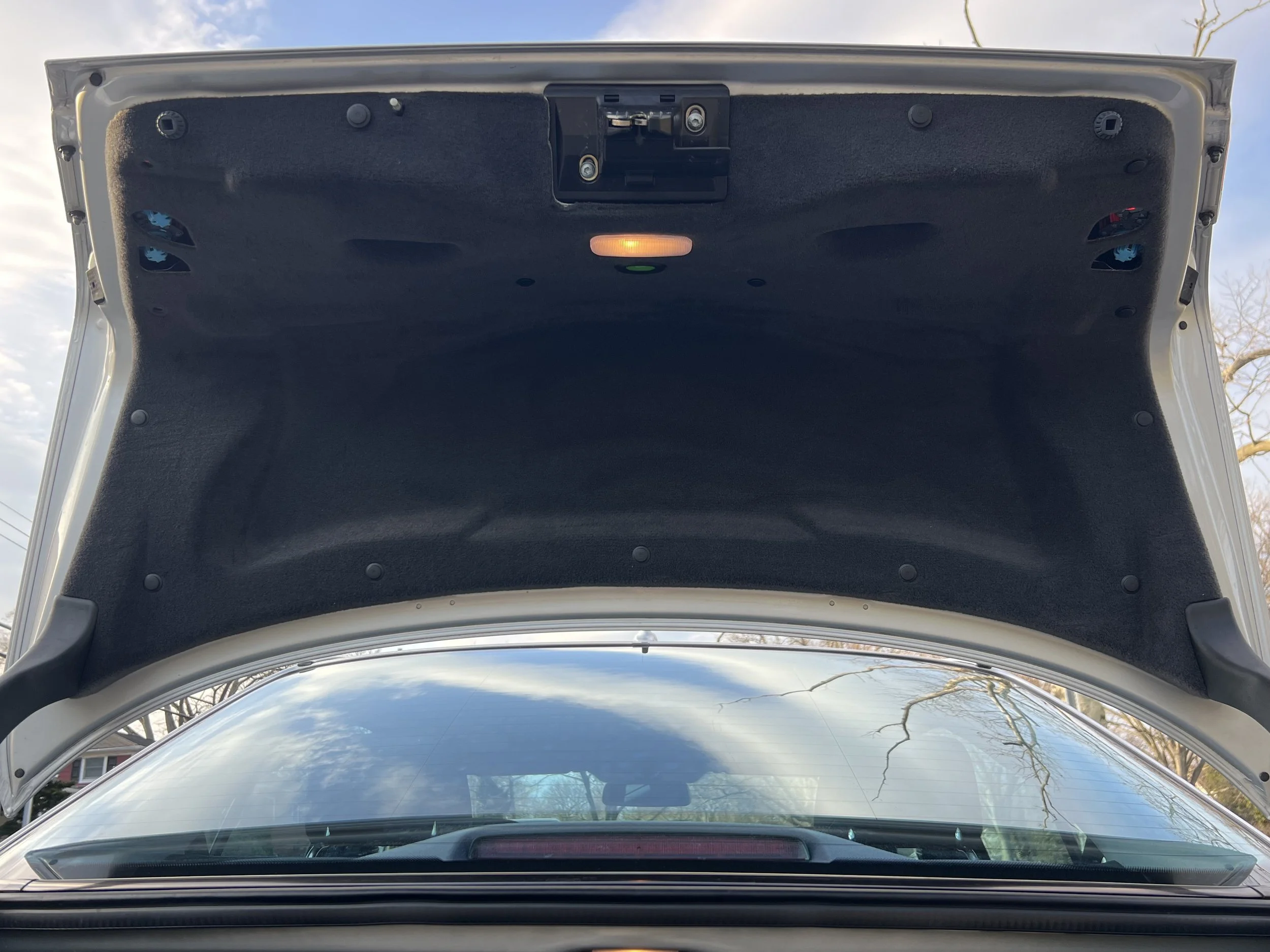 Open car trunk with interior lining and lights, reflecting a cloudy sky and trees on the windshield