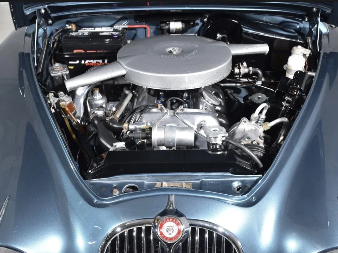 Close-up of a vintage car engine in a blue car with a prominent silver air filter cover and various engine components visible.