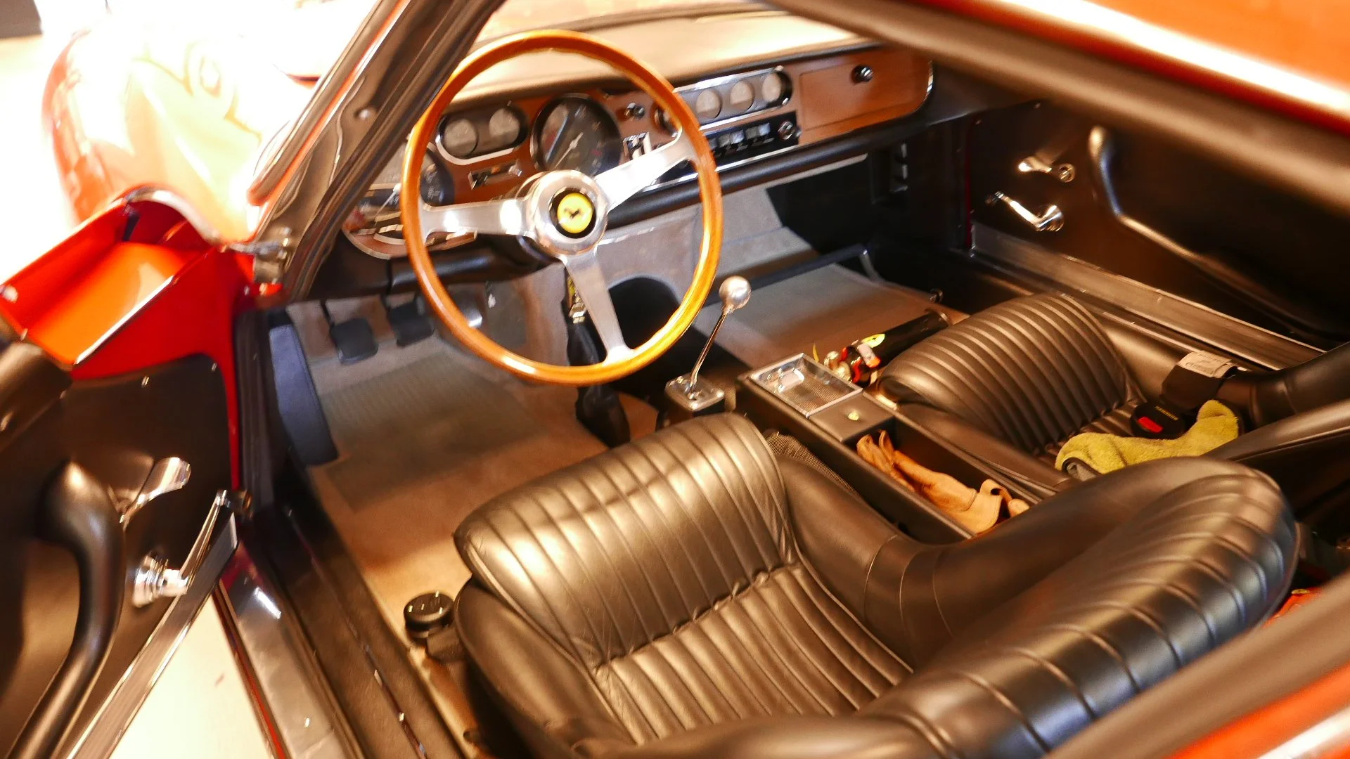 Interior of a classic Ferrari car, showing a wooden steering wheel, black leather seats, dashboard with gauges, gear shifter, and various control knobs.