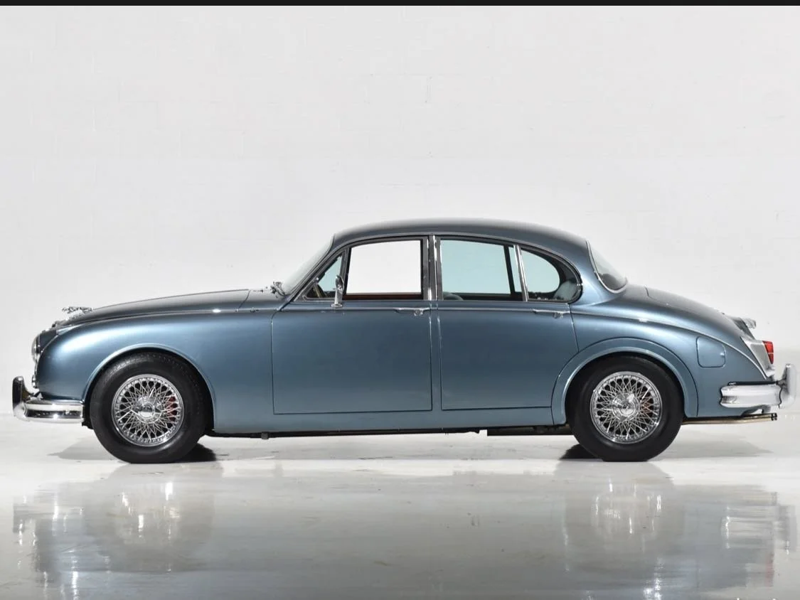 A classic vintage blue sedan car with wire-spoke wheels parked indoors against a white wall.