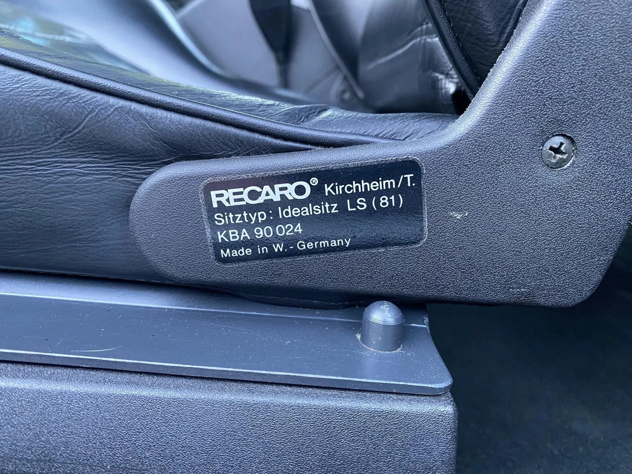 Close-up of a black label on a car seat with white lettering. The label reads "RECARO Kirchheim/T. Sitztyp: Idealsitz LS (81) KBA 90024 Made in W. Germany." The surrounding area shows black leather and metal components of the seat.