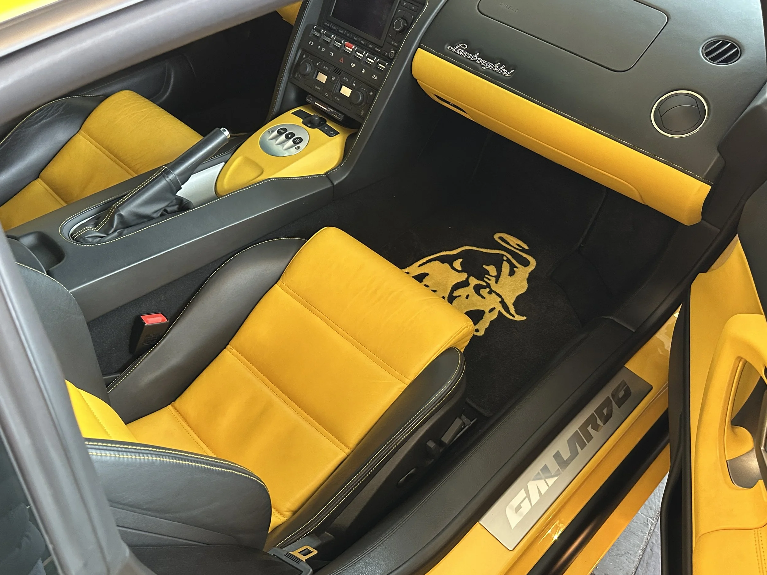 Interior of a yellow Lamborghini with yellow leather seats and black accents, showing the dashboard, steering wheel, and door sill with Lamborghini logo.
