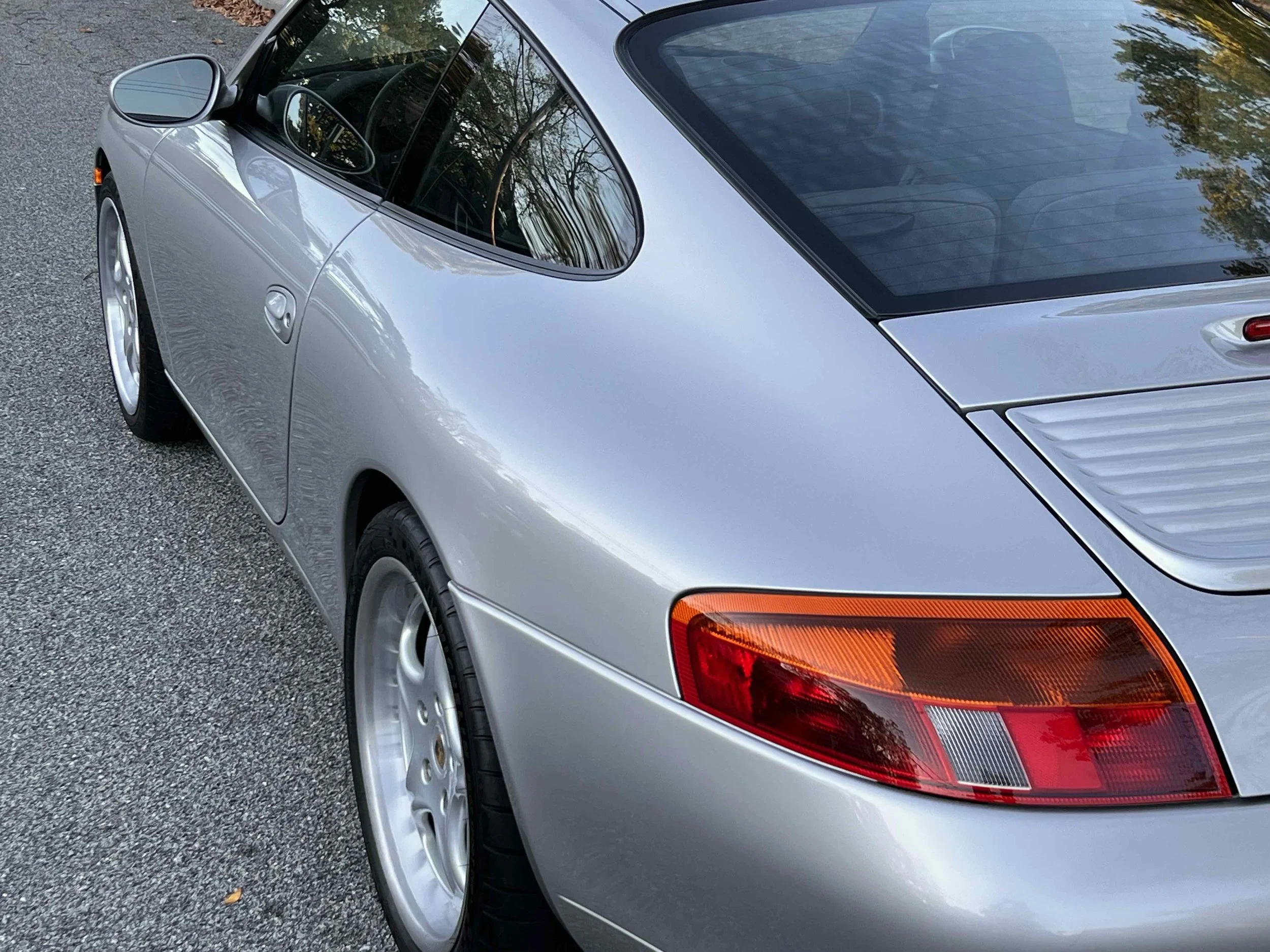 Silver classic Porsche 911 sports car parked on a paved surface, seen from the rear-left side with reflections of trees on the window