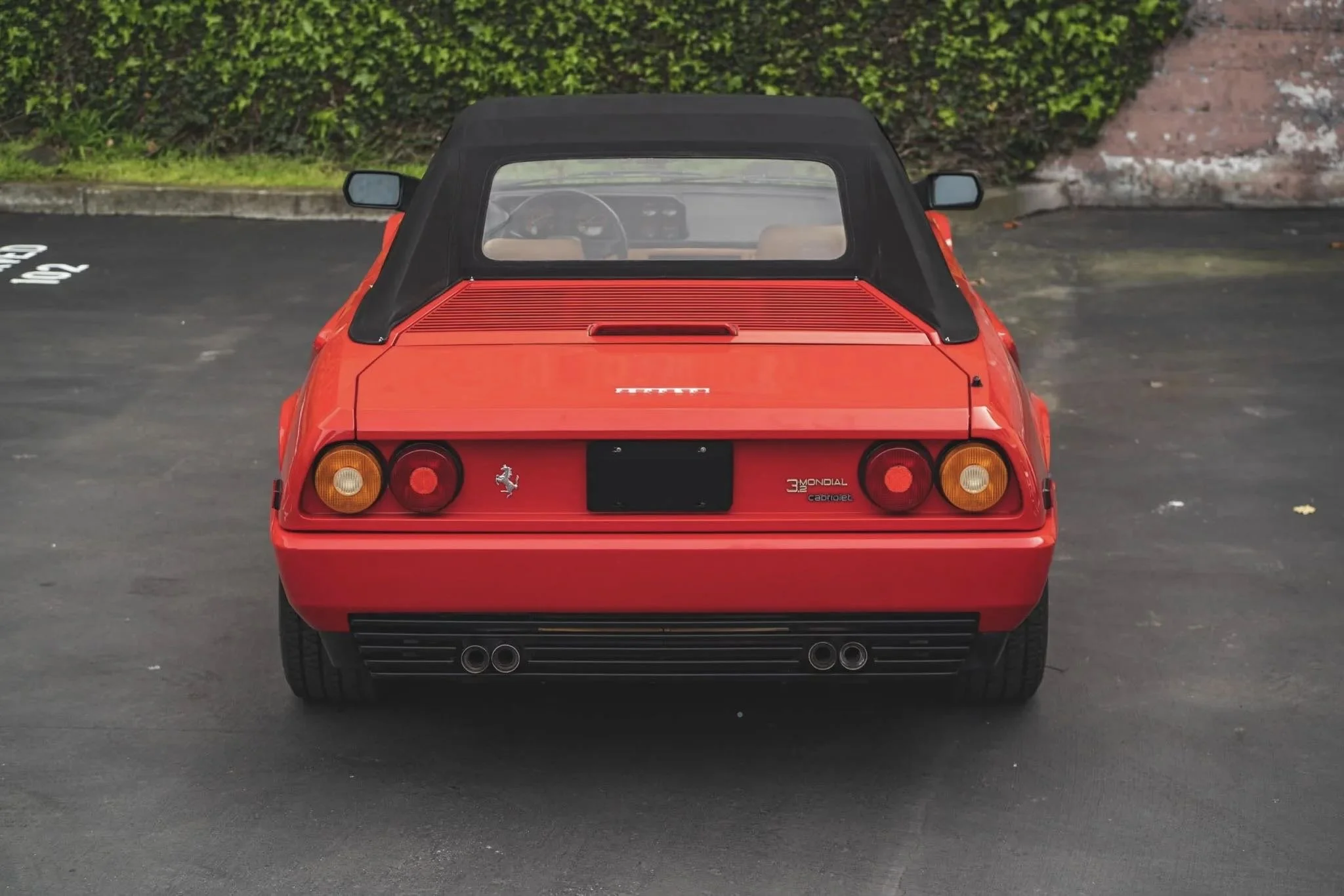 Red Ferrari 3.2 Mondial Cabriolet parked on a paved lot, showing its rear view with black soft top, round tail lights, and dual exhaust pipes.