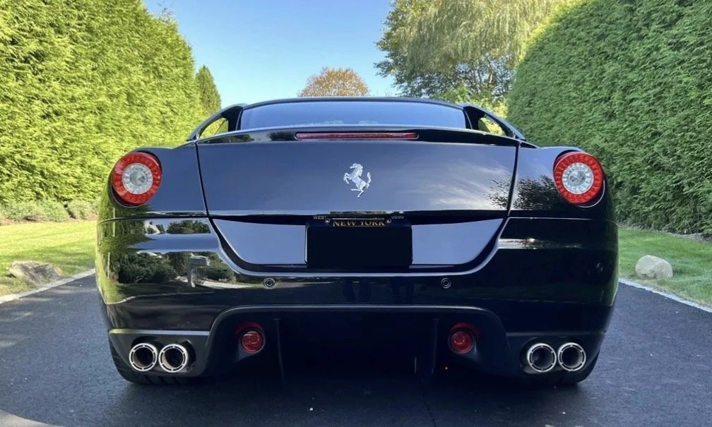 Back view of a black Ferrari sports car parked on a paved road with green bushes on both sides.