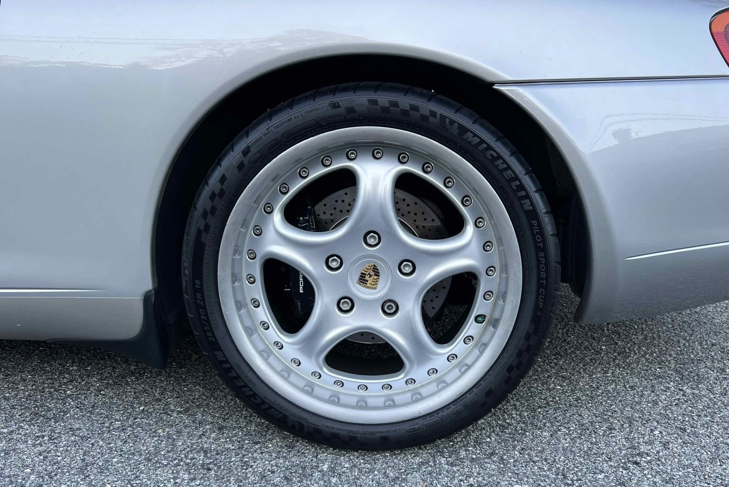Close-up of a silver Porsche car wheel with a Michelin Pilot Sport Cup 2 tire and a silver alloy rim featuring the Porsche crest in the center.