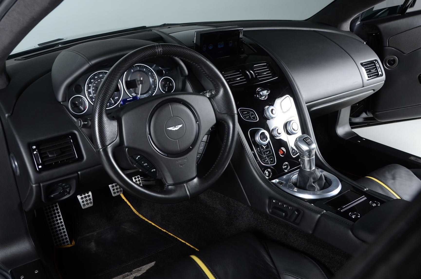 Inside view of a luxury sports car showing the steering wheel with a logo, the dashboard, center console with various controls, gear shift, and part of the driver's seat.