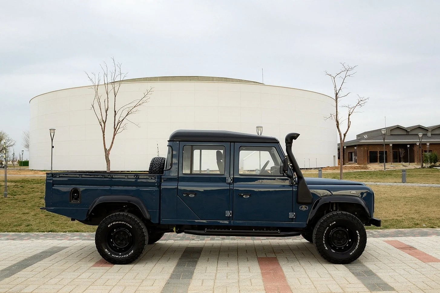 A dark blue pickup truck parked on a paved area with a large white circular building, leafless trees, grass, and a building in the background.