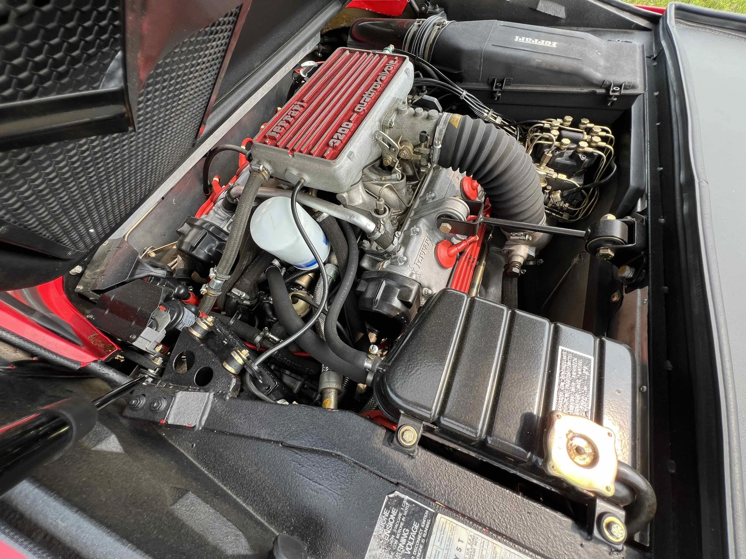 Close-up of a car engine bay featuring a red valve cover, black hoses, and various mechanical components.