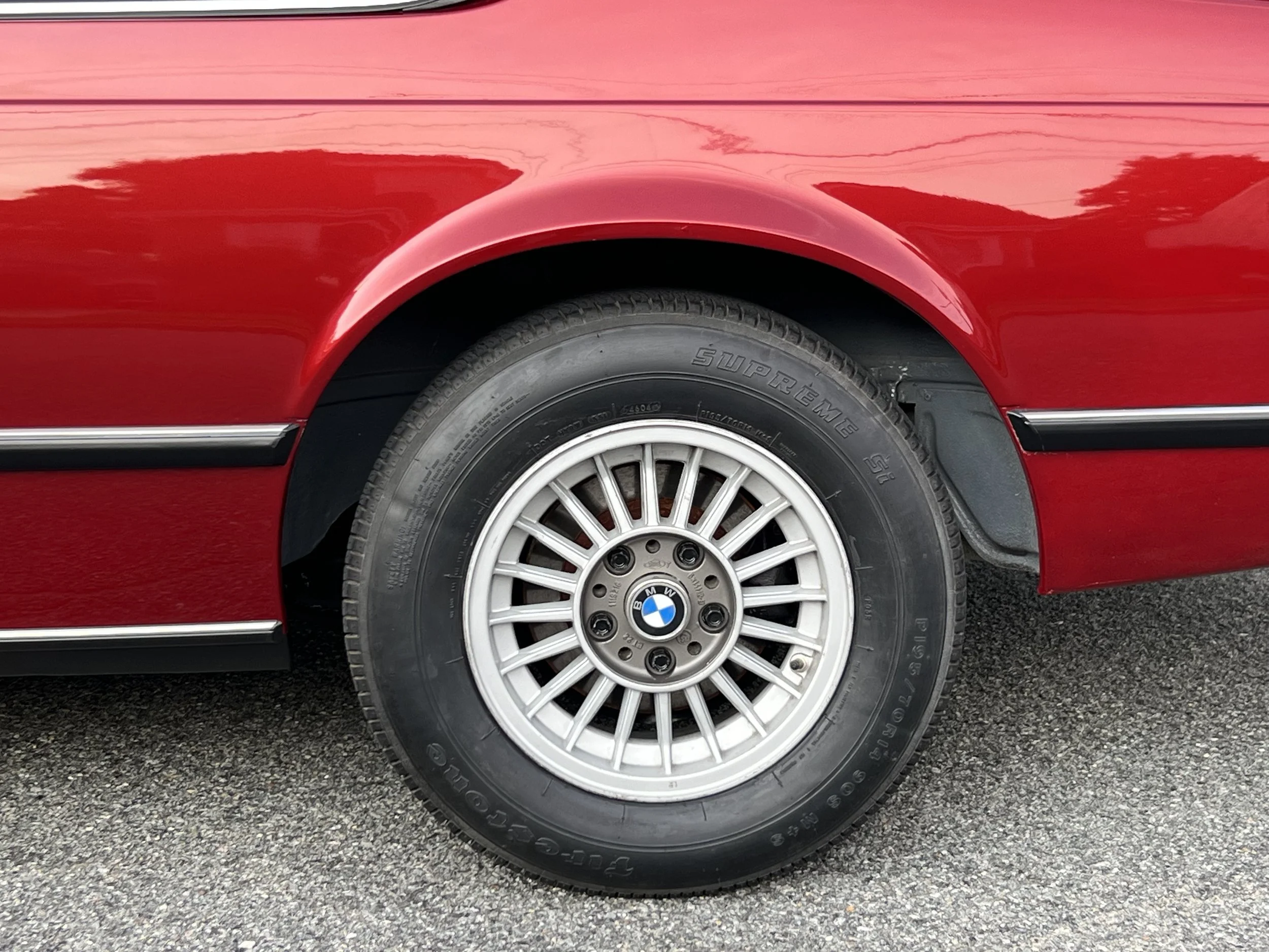 Close-up of a red BMW 630 csi front wheel with a BMW logo, tire brand 'Supreme SL', and a portion of the red car body on an asphalt surface.