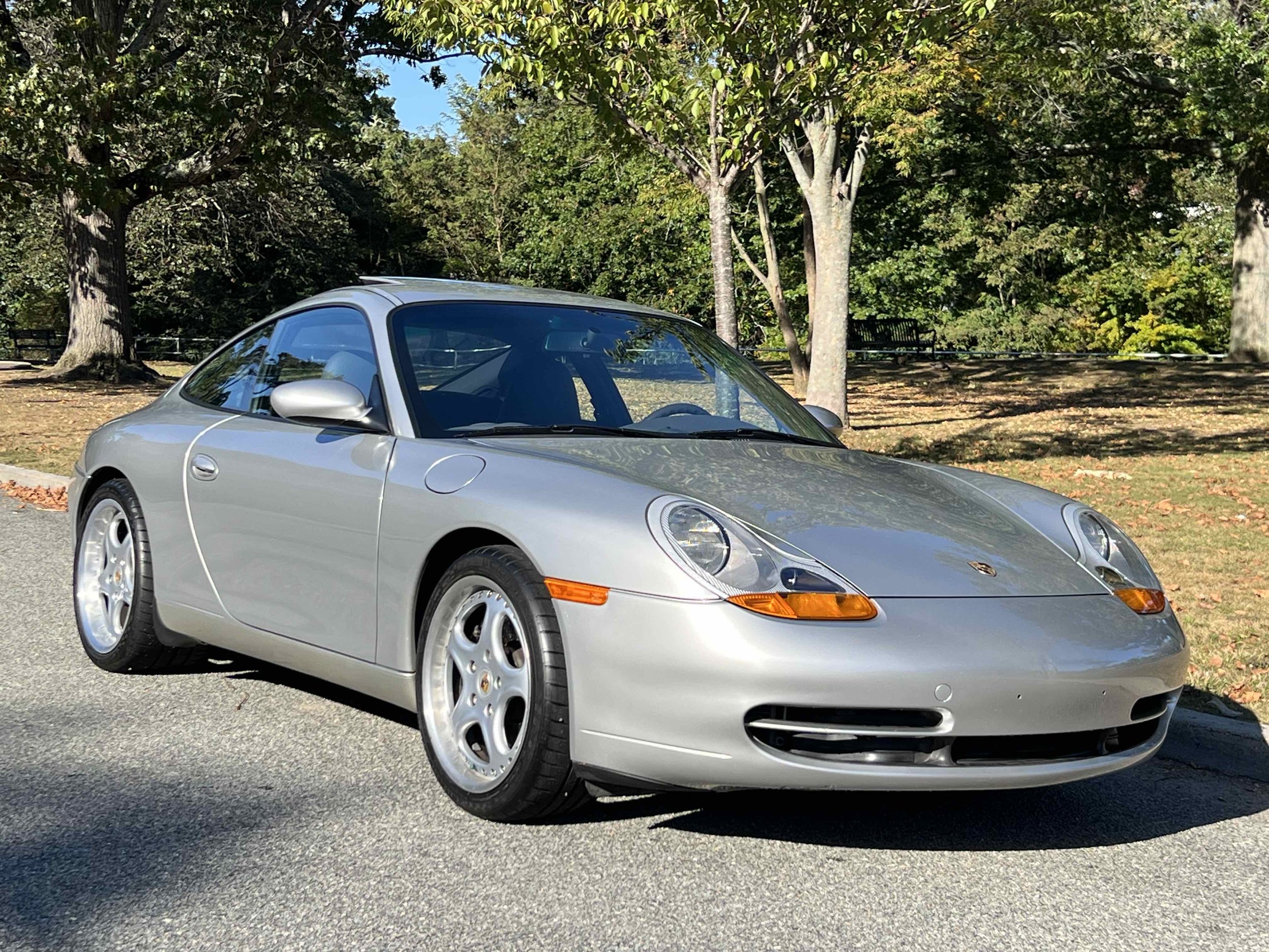 Silver Porsche 911 sports car parked on a road with trees and grass in the background.