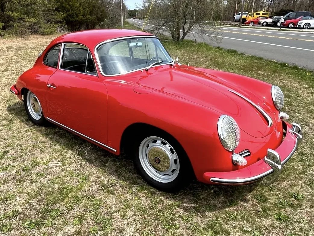 Red vintage Porsche 356 classic car parked on grassy area near road with trees and other vehicles in background