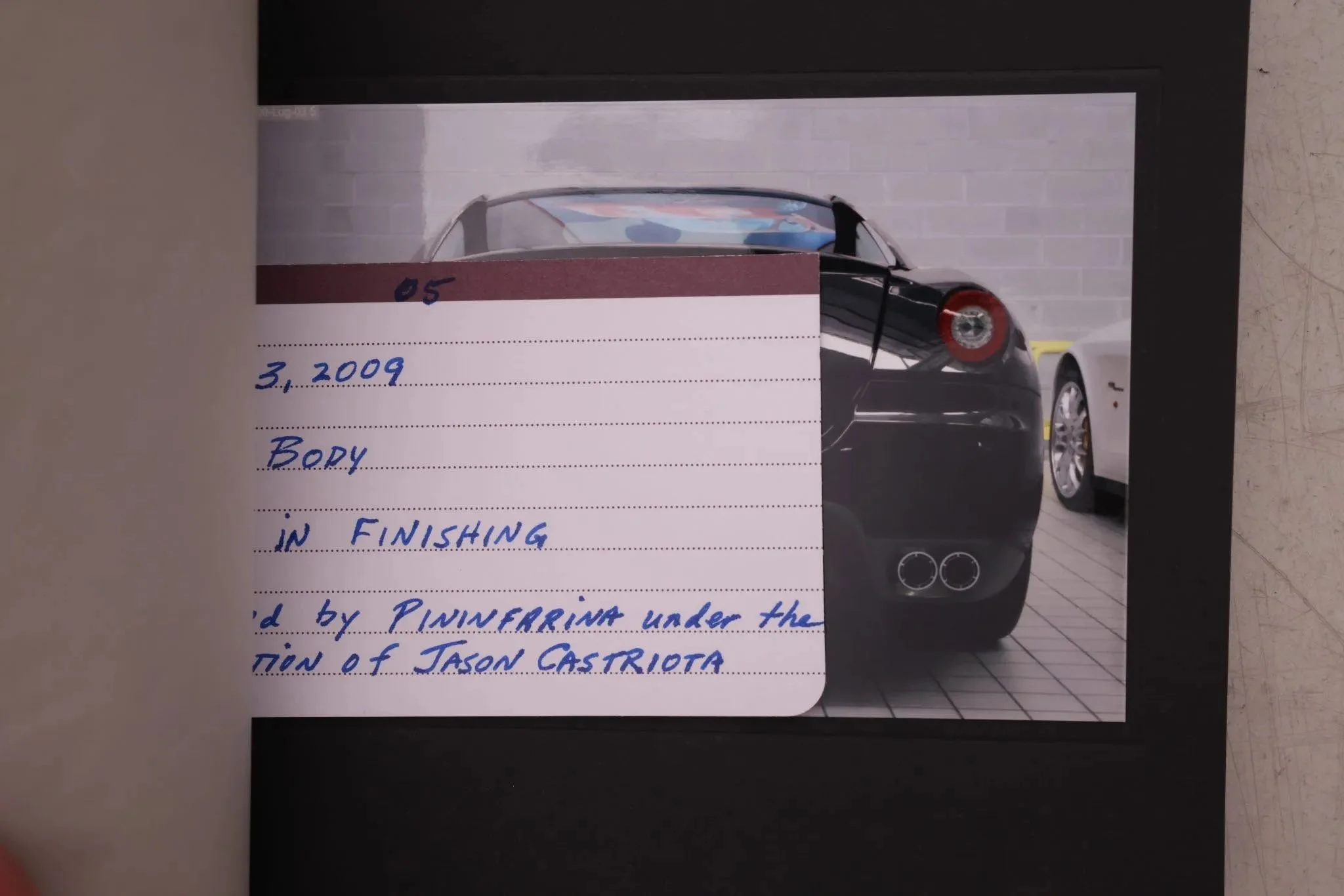 Photograph of a black sports car with dual exhaust pipes, parked in a garage with a white wall and a second white car next to it. There is a handwritten note overlaying the image, partially obscuring the view of the black car.
