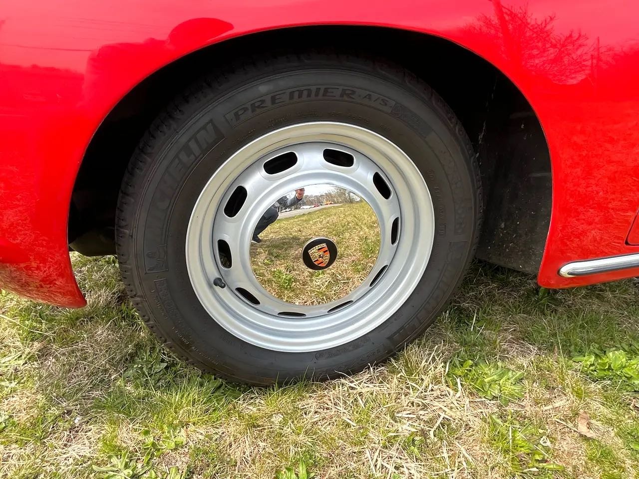 Close-up of a red car's wheel with a silver hubcap featuring a Porsche logo, on grass, with a reflection of a person taking the photo visible in the hubcap