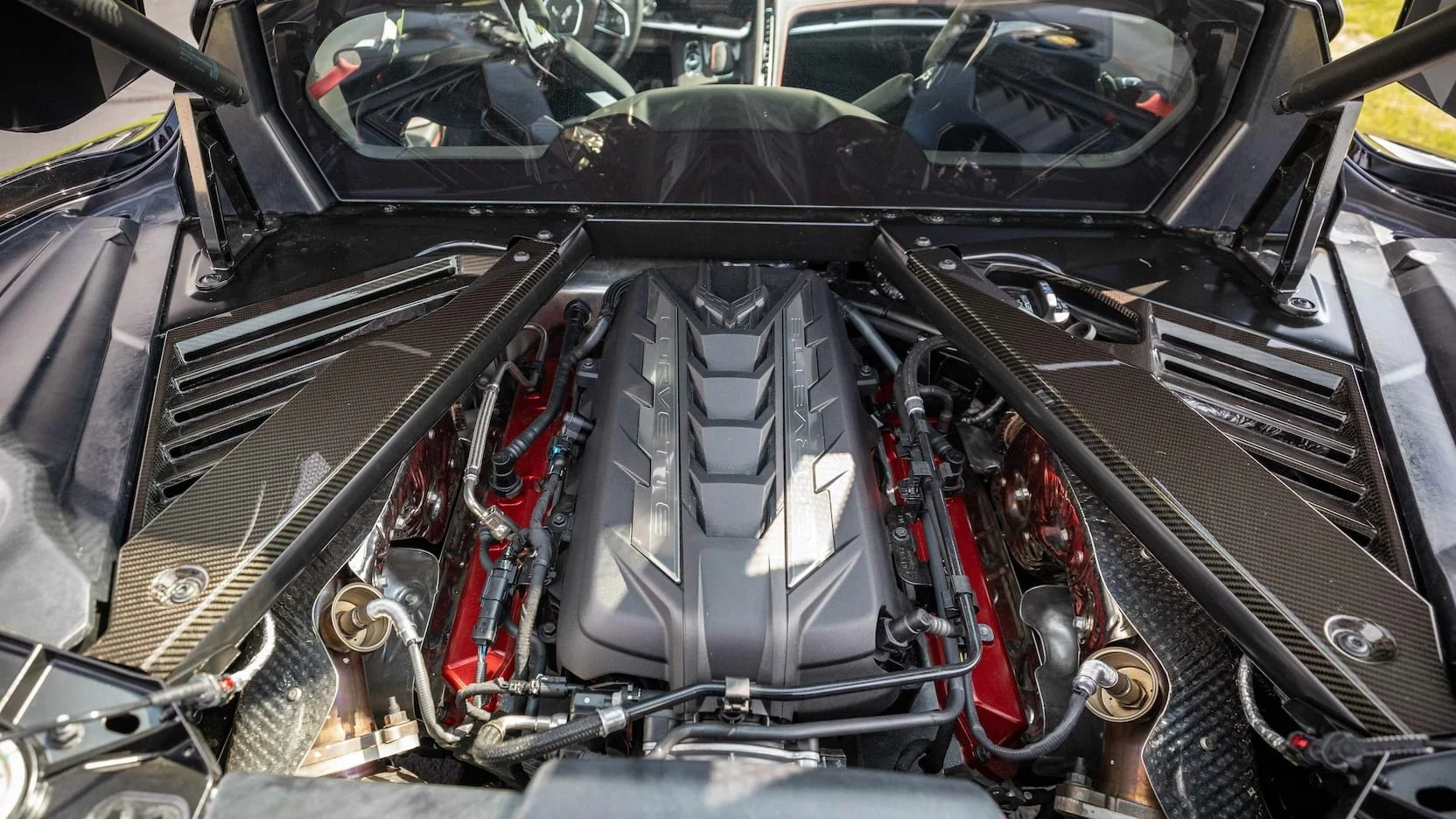 A supercar engine bay featuring a central V12 engine with a black cover, surrounded by carbon fiber panels and various mechanical and electrical components.