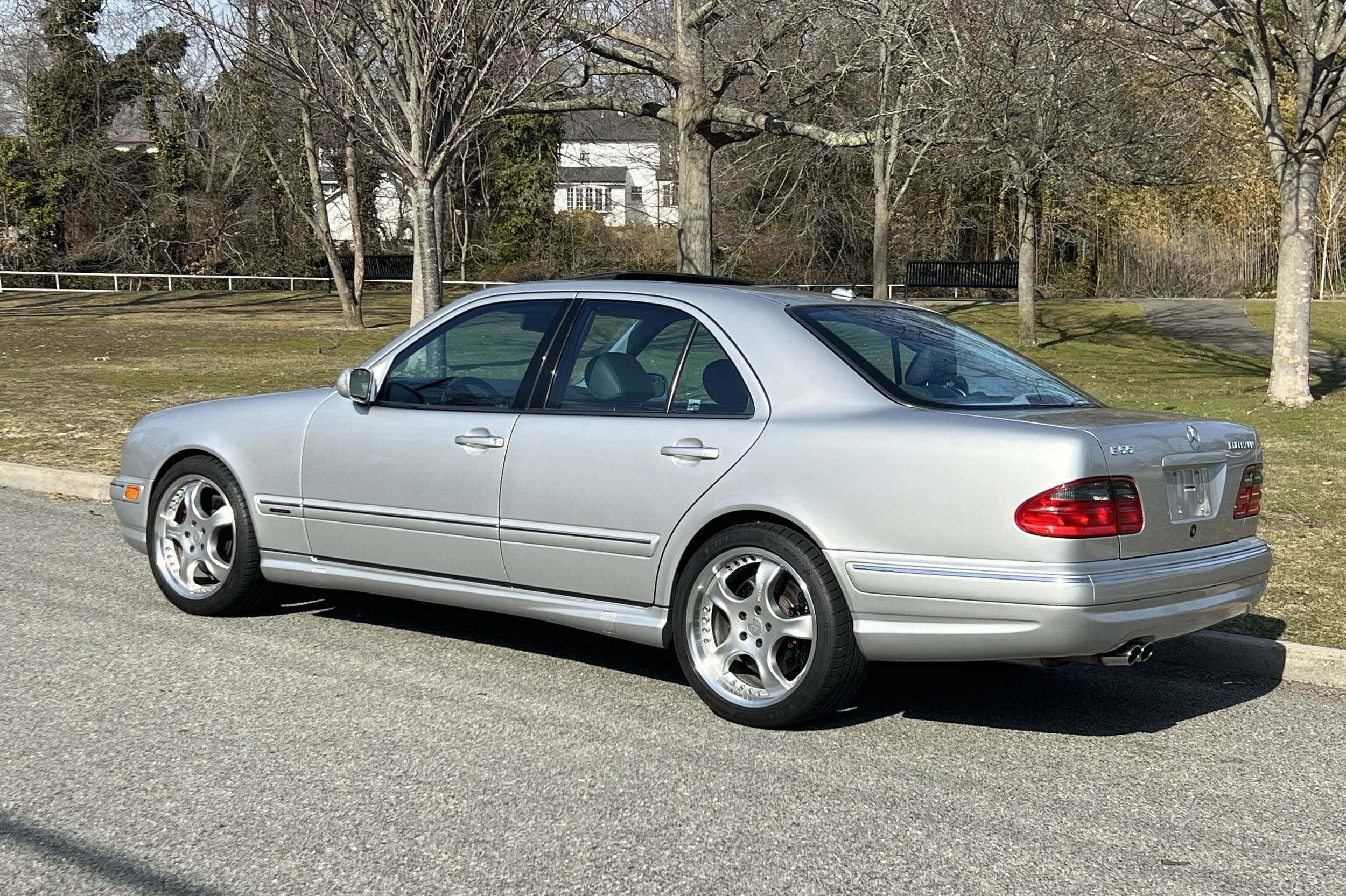 A silver Mercedes-Benz sedan parked on the side of a street near a park with trees and benches.