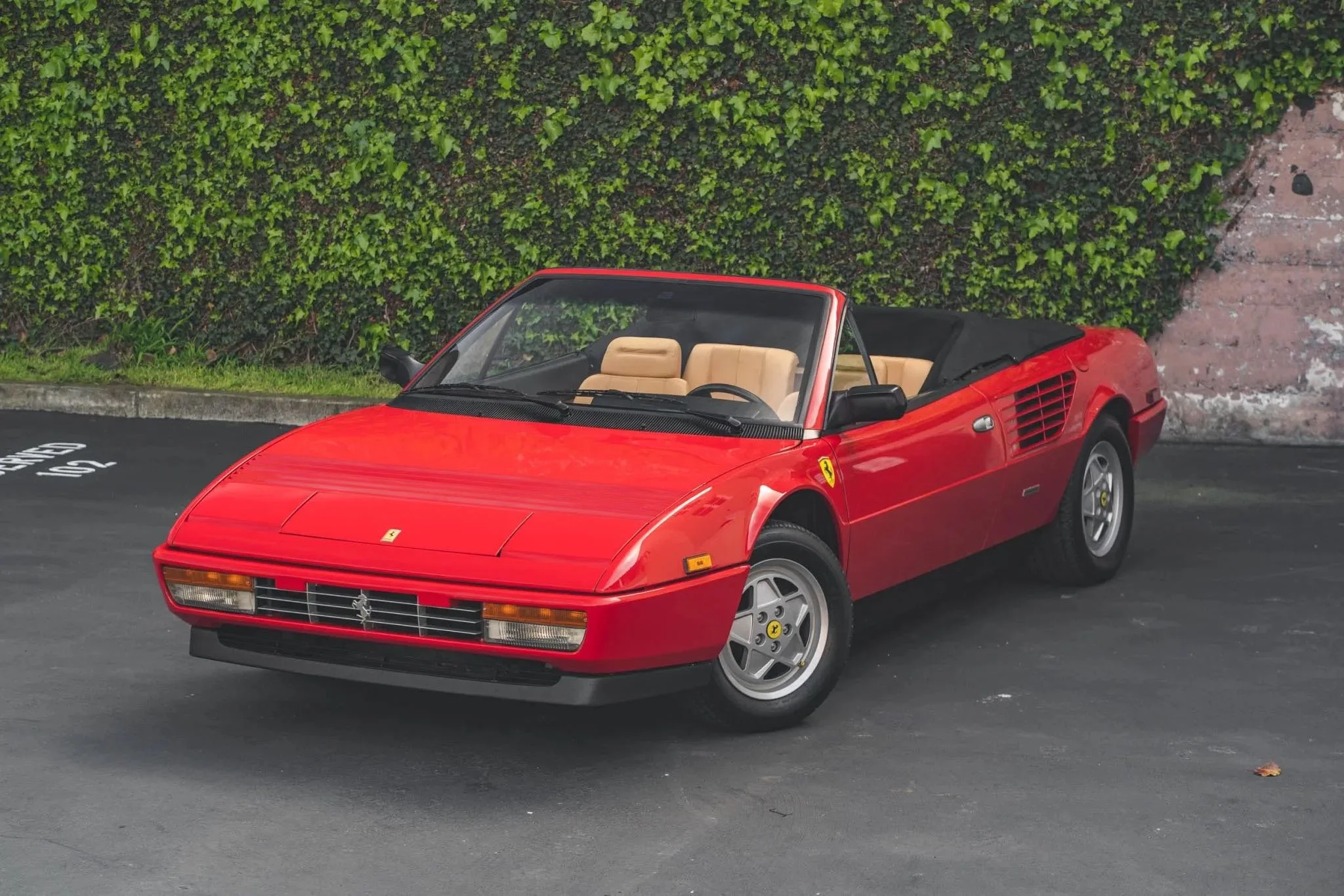 A red vintage Ferrari convertible car parked on a paved surface with green leafy bushes in the background.