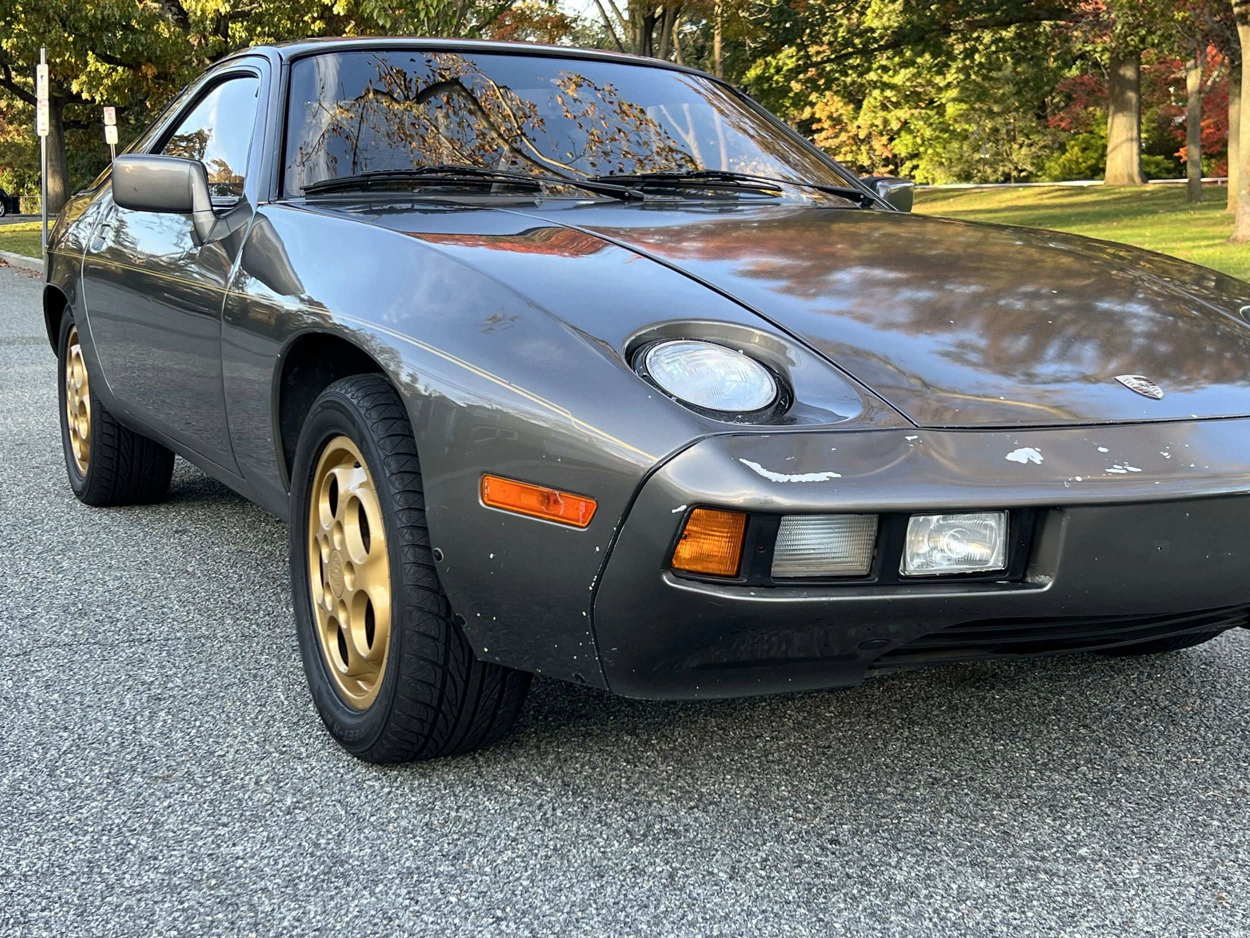 A vintage gray Porsche sports car with gold wheels parked on a street with sequoia trees in the background.