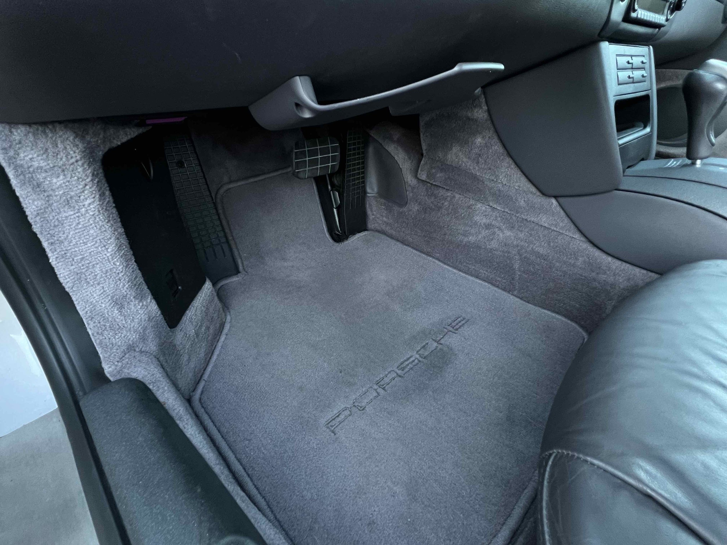 The front passenger footwell of a car with a gray carpeted floor mat, black pedals, and interior panels, and a portion of a leather seat visible in the bottom right corner.