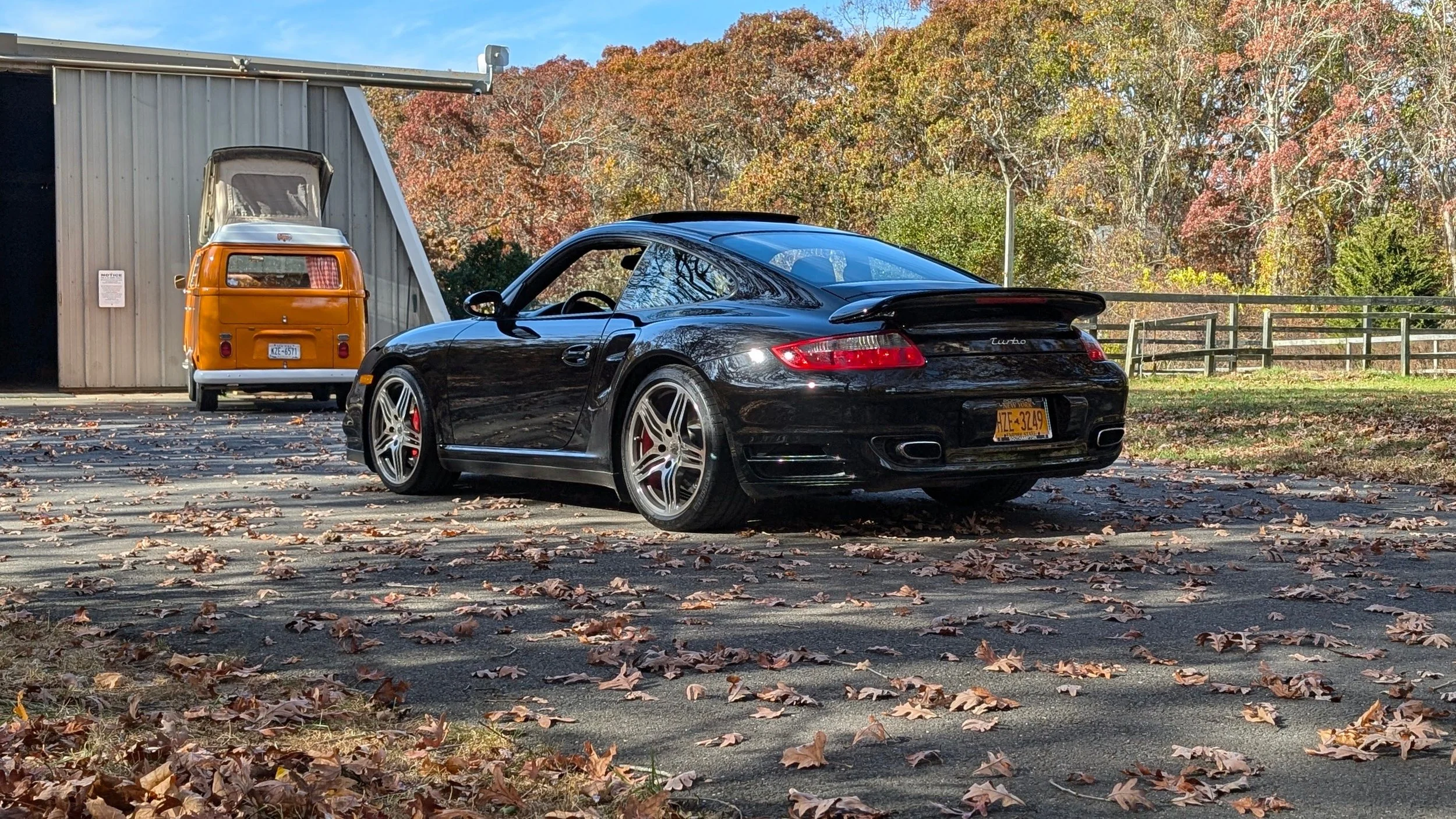 A black Porsche sports car parked outdoors with a black spoiler, colorful autumn leaves on the ground, and a vintage orange and white van with a pop-up roof parked near a gray industrial building against a backdrop of trees with fall foliage.