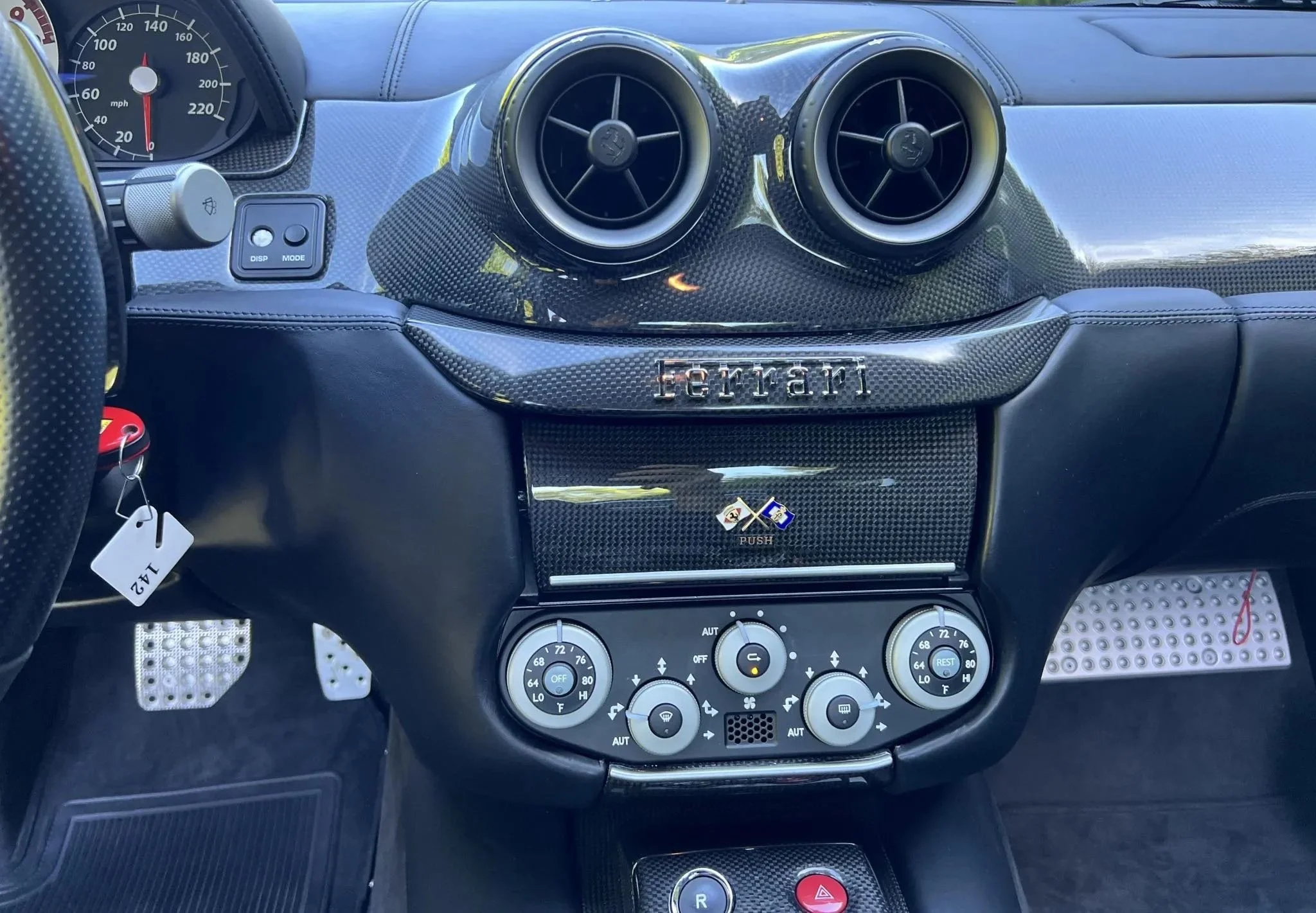 The interior dashboard of a Ferrari sports car with carbon fiber accents, circular air vents, and climate control system.