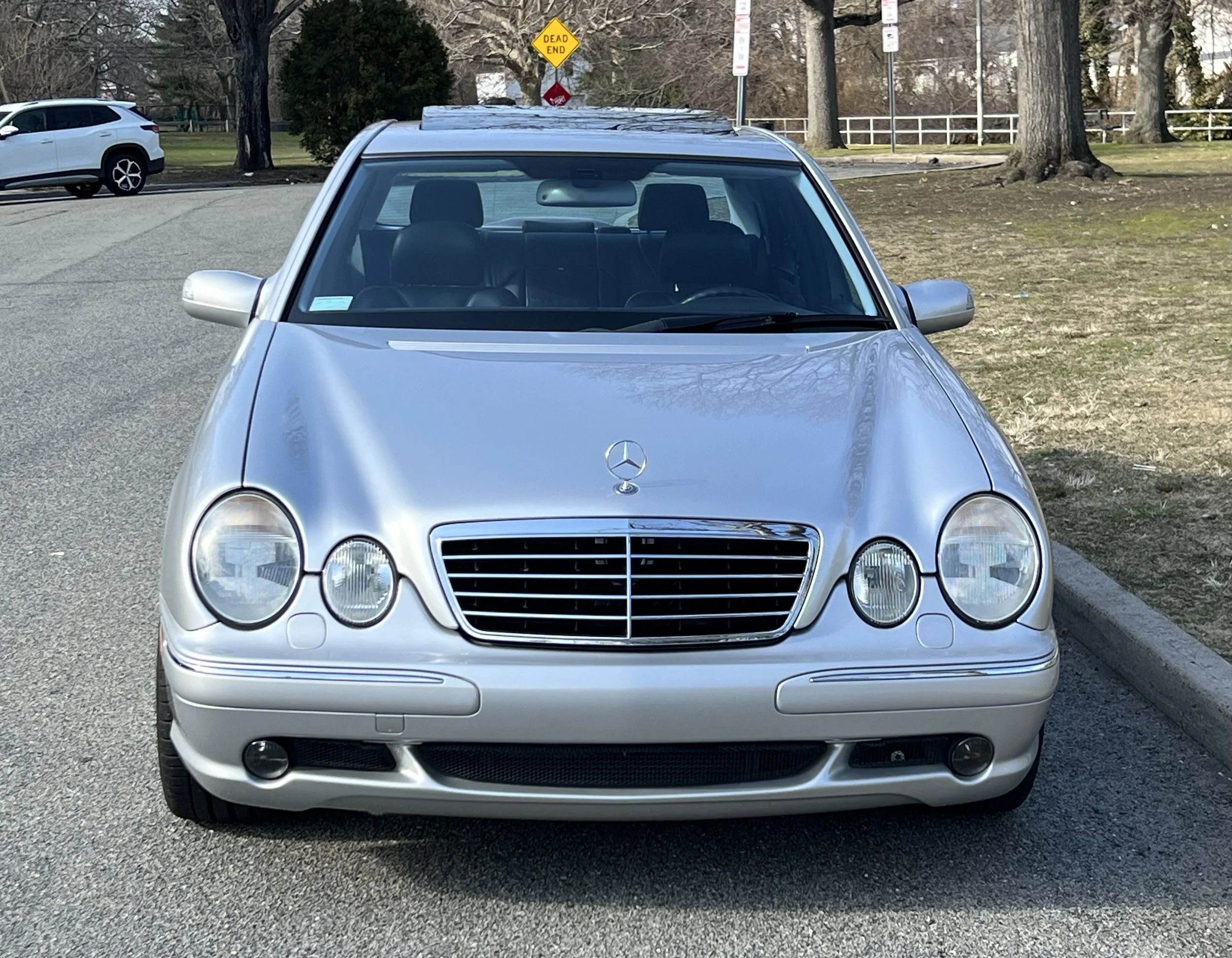 Front view of a silver Mercedes-Benz car parked on a street, with a white SUV in the background and a yellow 'Dead End' street sign visible behind the Mercedes.
