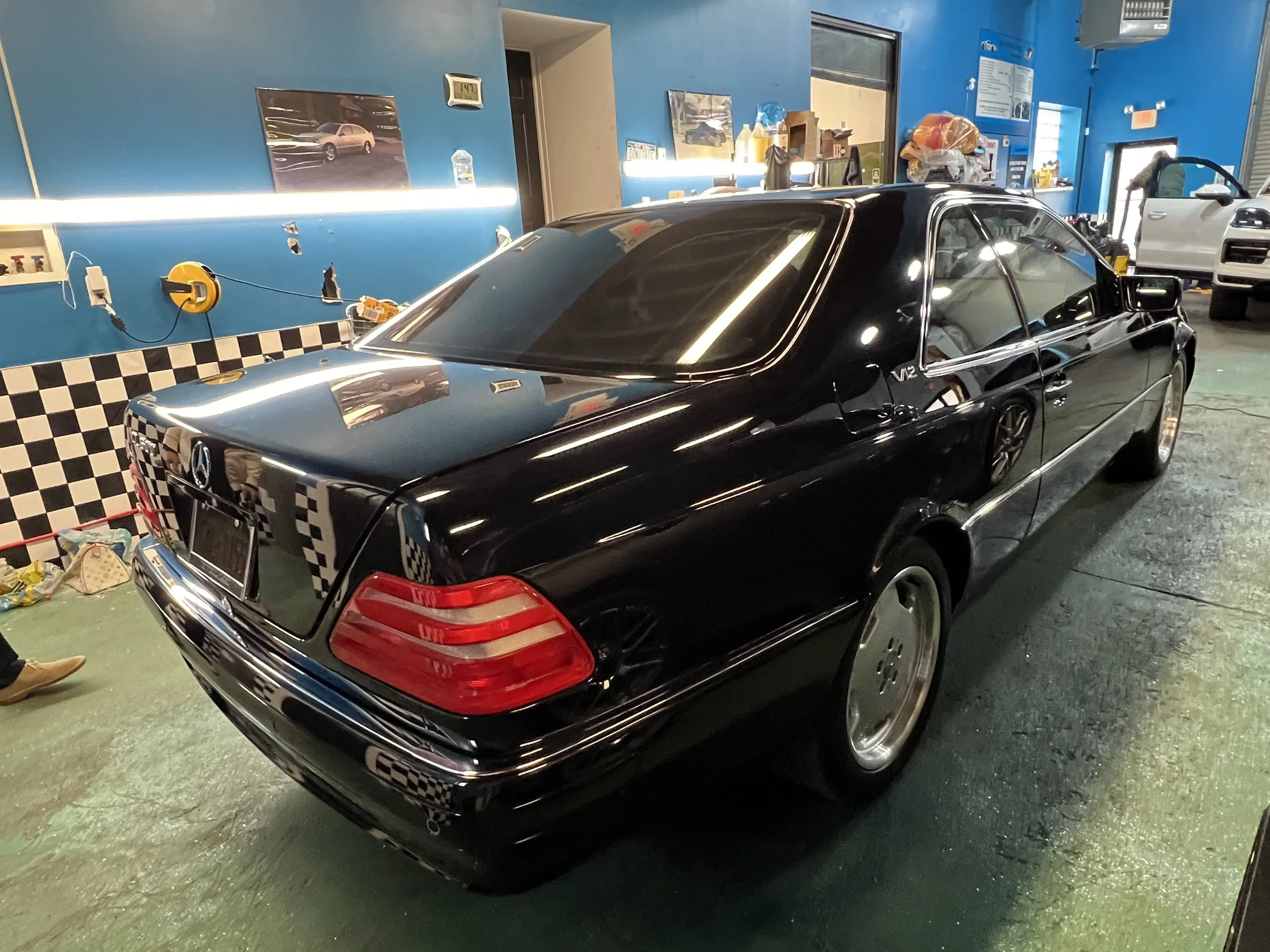 Black vintage Mercedes-Benz car inside a garage with blue walls, checkered black-and-white wallpaper, and various tools and objects around.