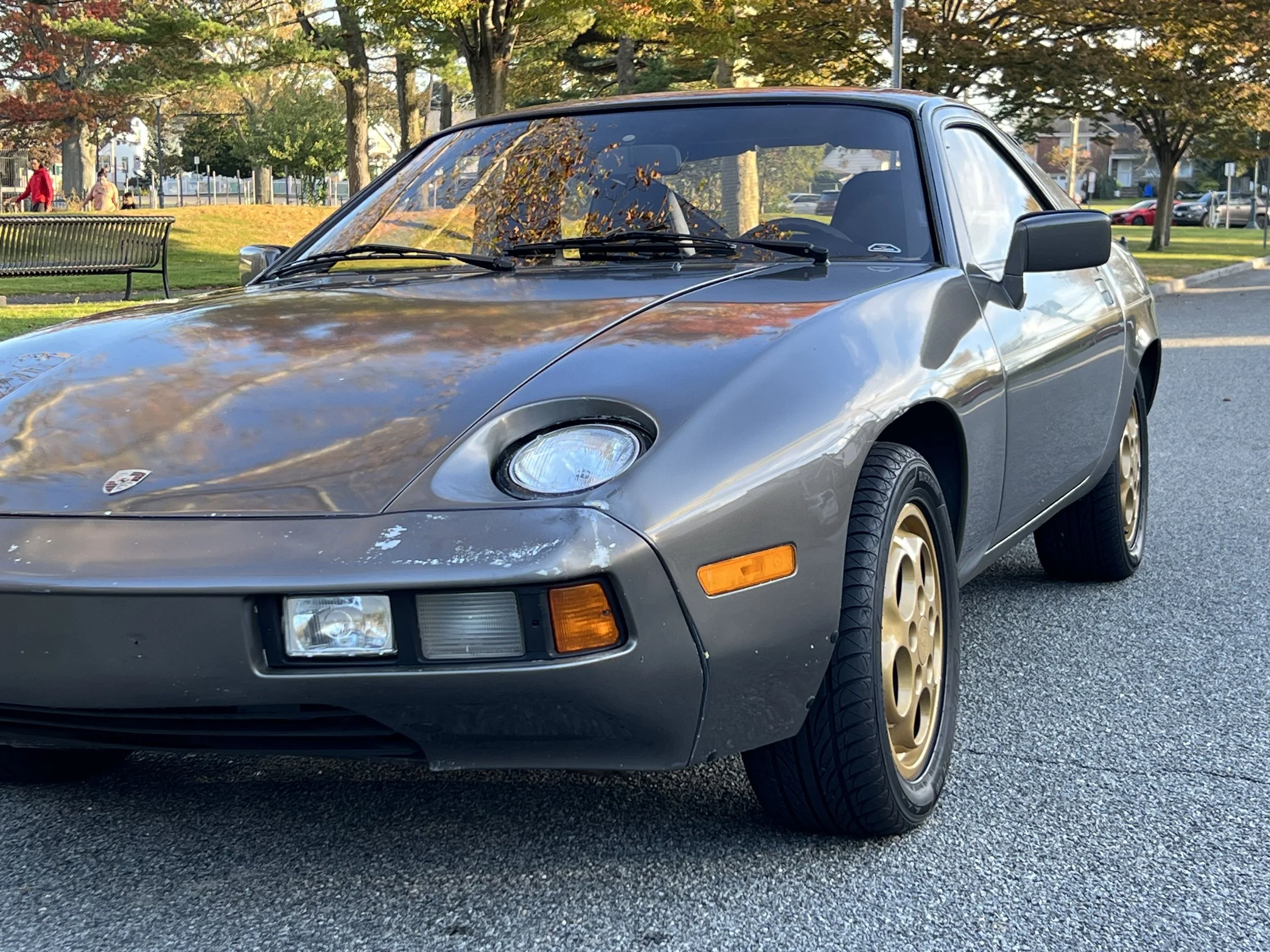 A vintage gray Porsche 944 sports car with gold wheels parked on a paved street in a park with trees, grass, benches, and people in the background.