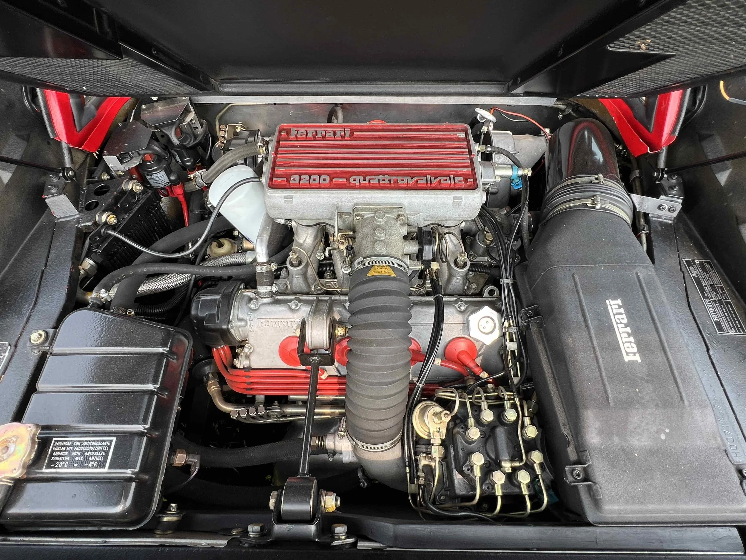 Engine bay of a Ferrari race car with a 3.0-liter V8 engine, featuring a red valve cover, black air intake, and various hoses and components.