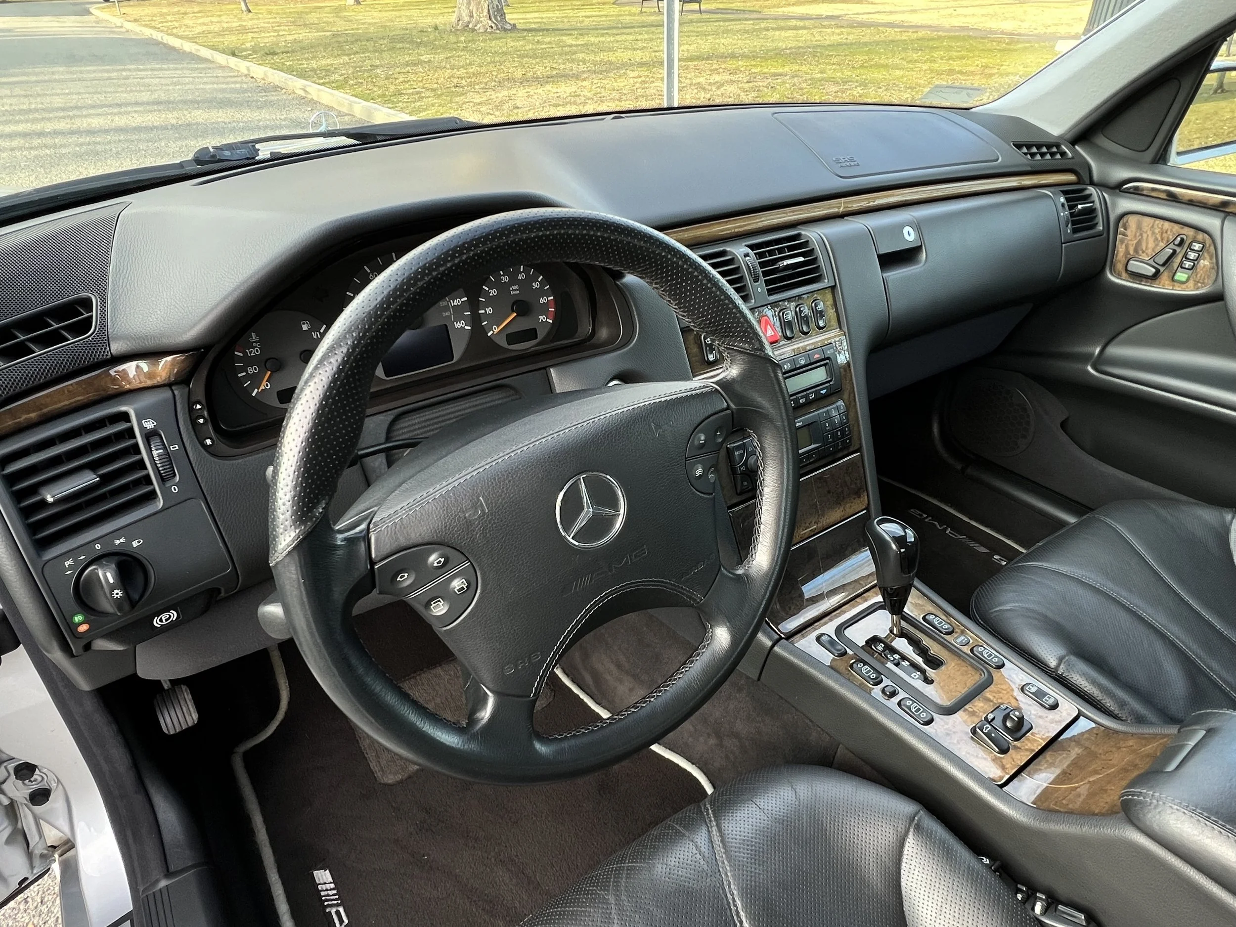 The interior of a Mercedes-Benz vehicle showing the steering wheel, instrument cluster, center console with controls, and part of the passenger seat, with a parking lot and grass visible through the windshield.