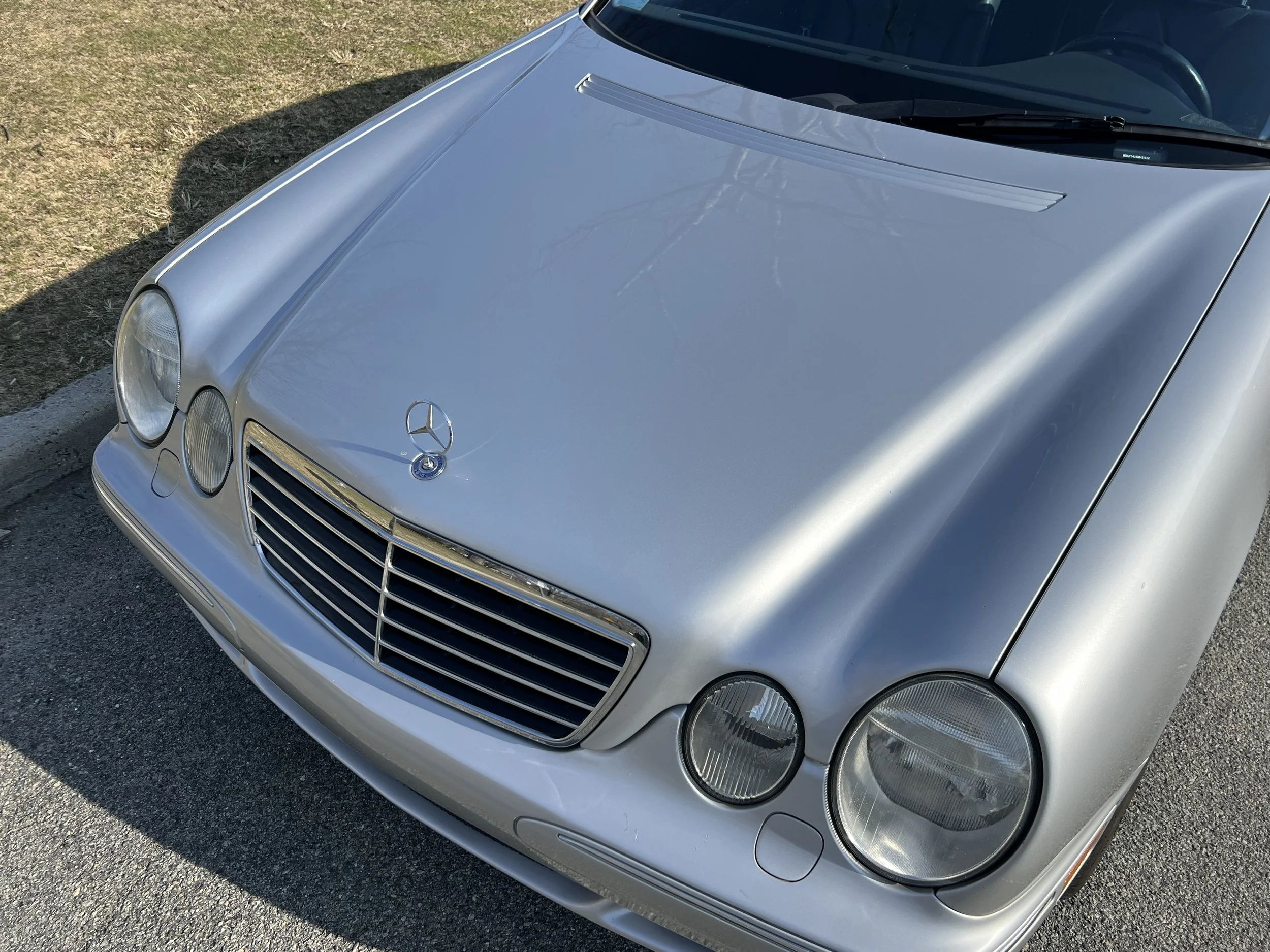 A silver Mercedes-Benz car parked on a gravel surface, showing the front left side including the headlight, grille, hood ornament, and windshield.