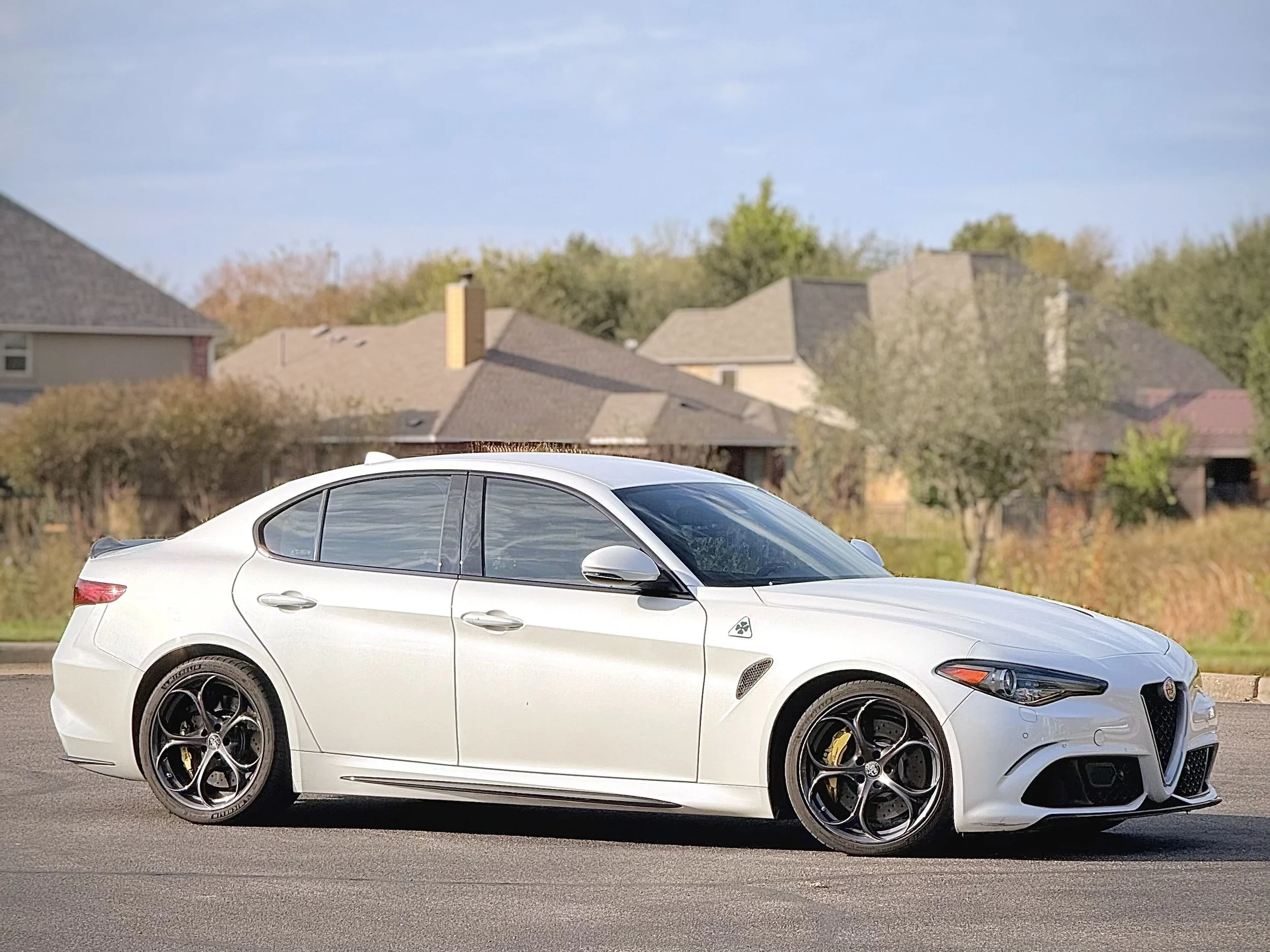 A white Alfa Romeo sedan parked on a residential street with houses and trees in the background.
