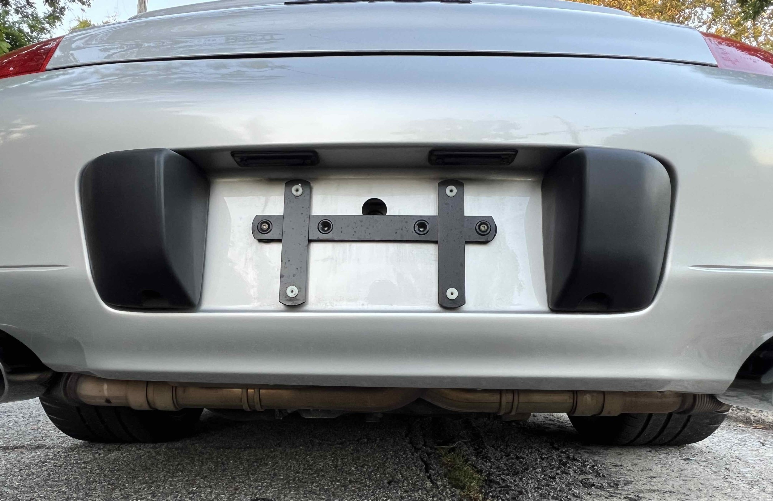 Rear of a silver car with the license plate removed, showing the mounting bracket for the license plate holder, with black mounting hardware and black fog lights on either side, and a visible exhaust pipe below.