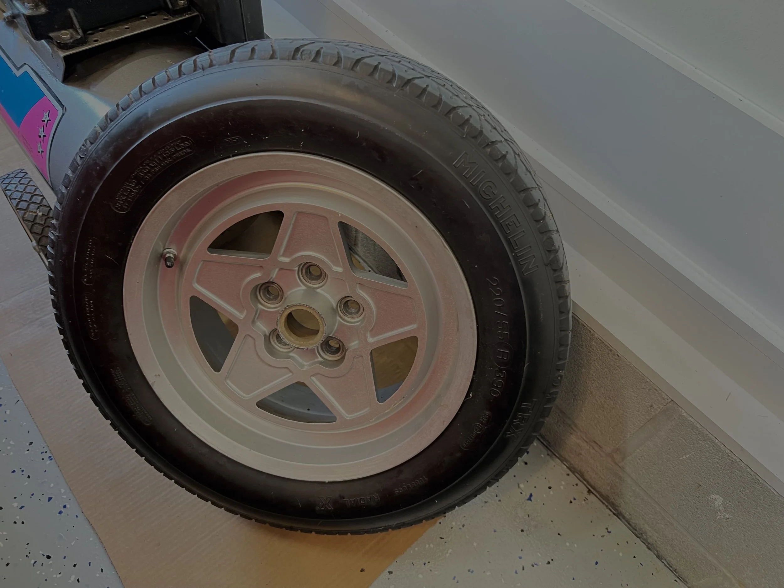 Close-up of a black tire mounted on a pink and white wheel, leaning against a garage wall.