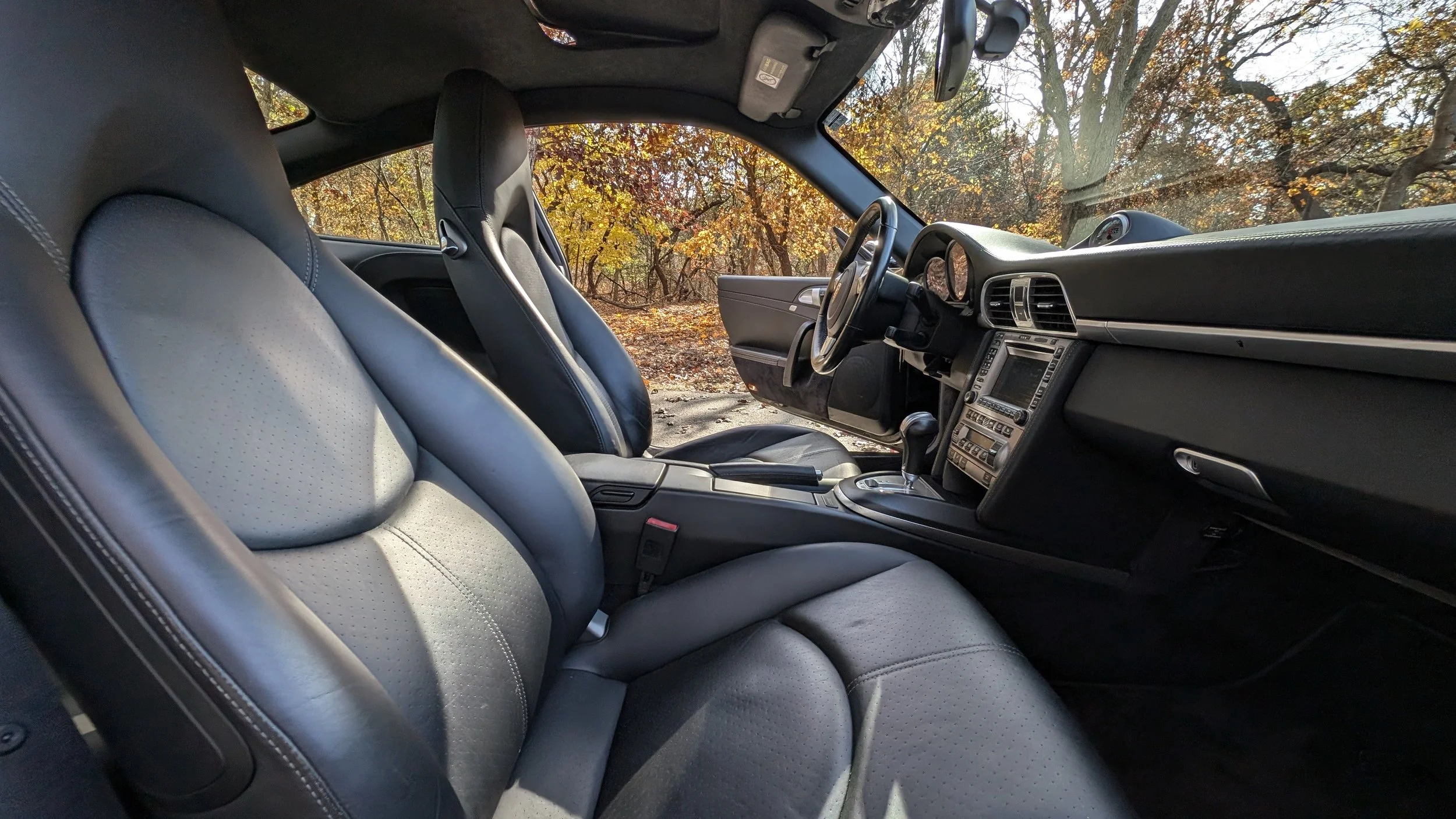 Interior view of a car showing two black leather seats, dashboard, gear shift, and a window with a view of trees with autumn leaves.