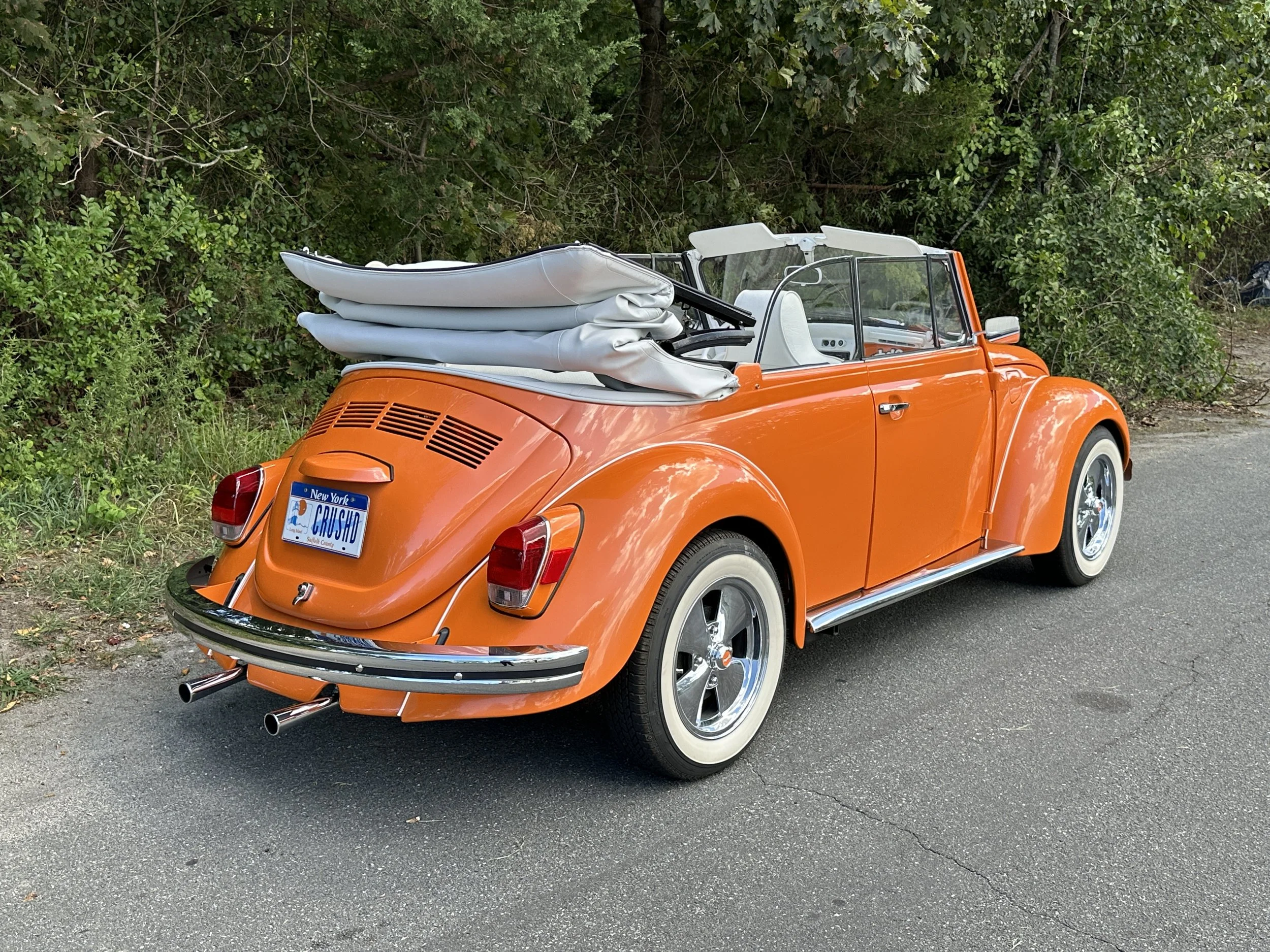 Orange vintage Volkswagen Beetle with white-wall tires, a roof rack carrying folded white tents, and a New York license plate that reads 'CRUSHD' parked on the side of a road next to green bushes.