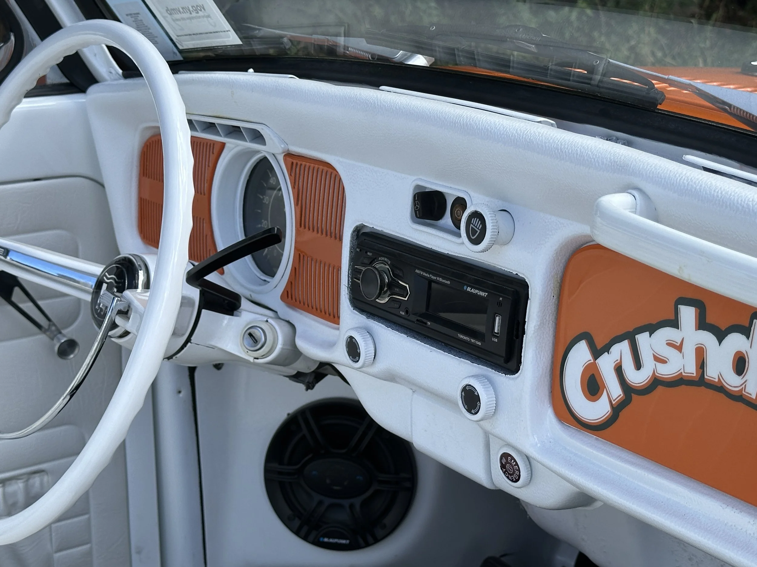 The interior of a vintage Volkswagen van with a white dashboard, orange accents, and a 'Crush' sticker on the dashboard.