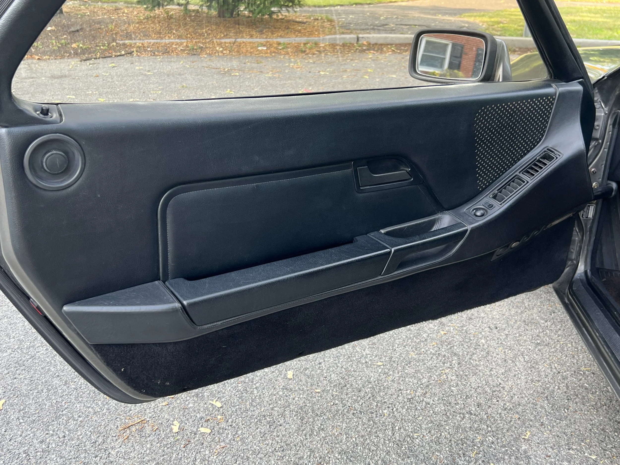 Close-up of a vintage black car door interior, showing a speaker, window controls, door handle, and storage compartment, with a parking lot in the background.