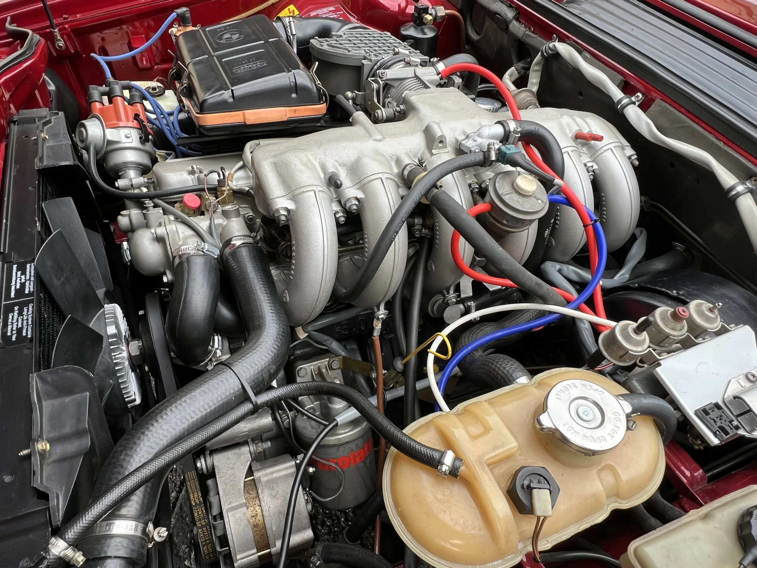 Close-up of a 1977 BMW 630 csi car engine bay showing various engine components, hoses, wires, and a turbocharger, with a red-painted interior.