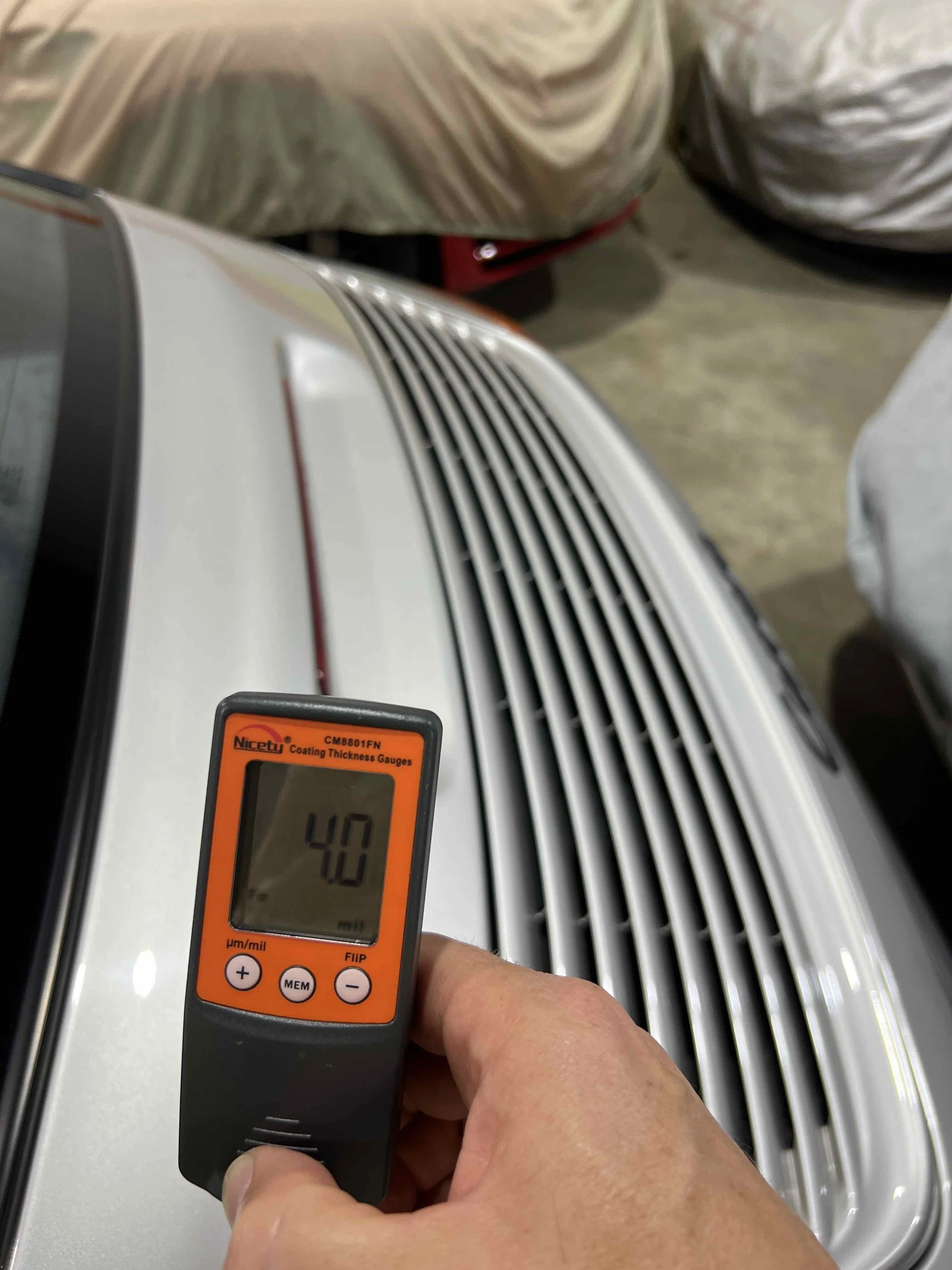 A coating thickness gauge measuring 40 micrometers being held in front of a white vehicle with a vented hood in a garage or workshop.