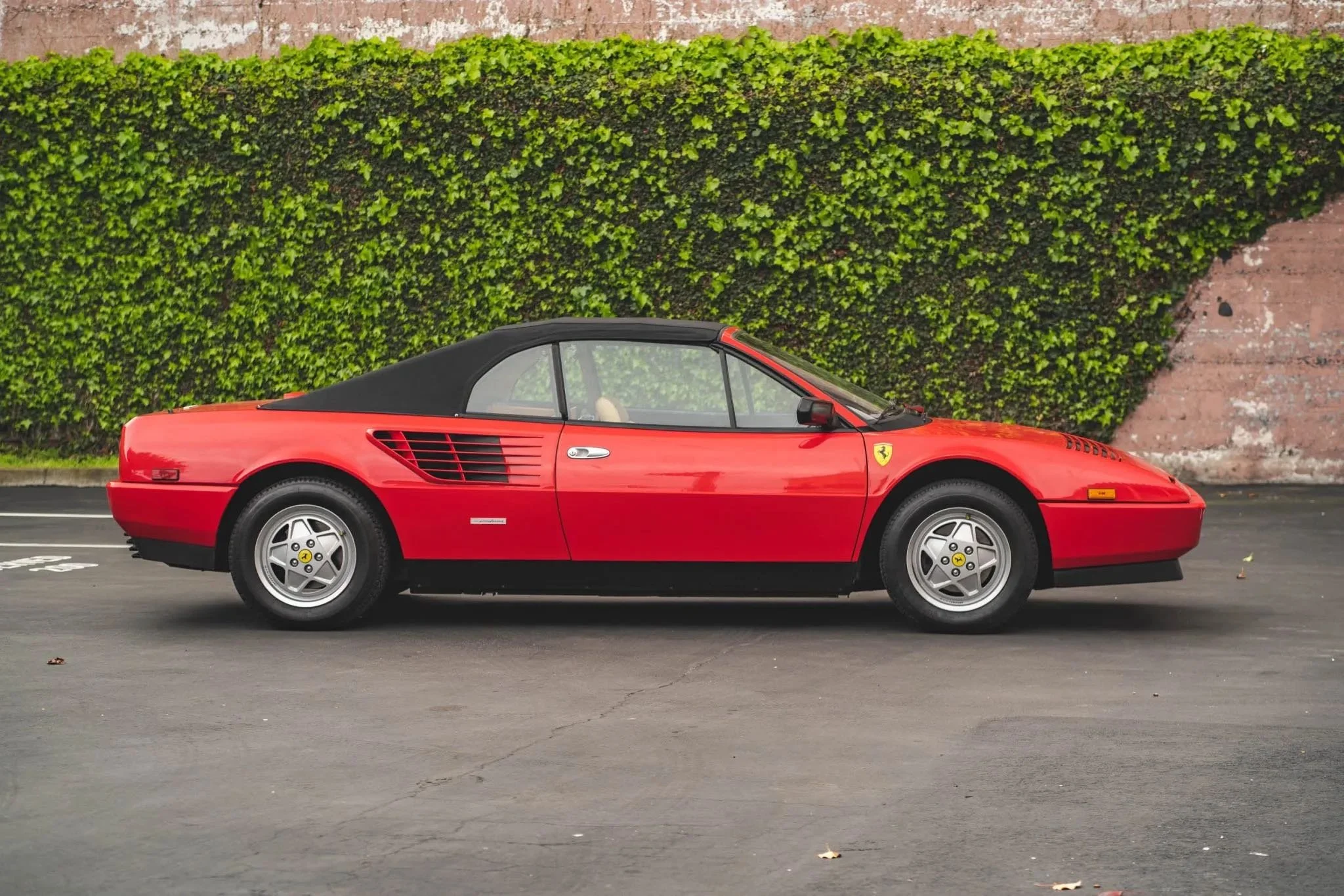 Red Ferrari sports car with black convertible top parked on a paved road in front of green bushes