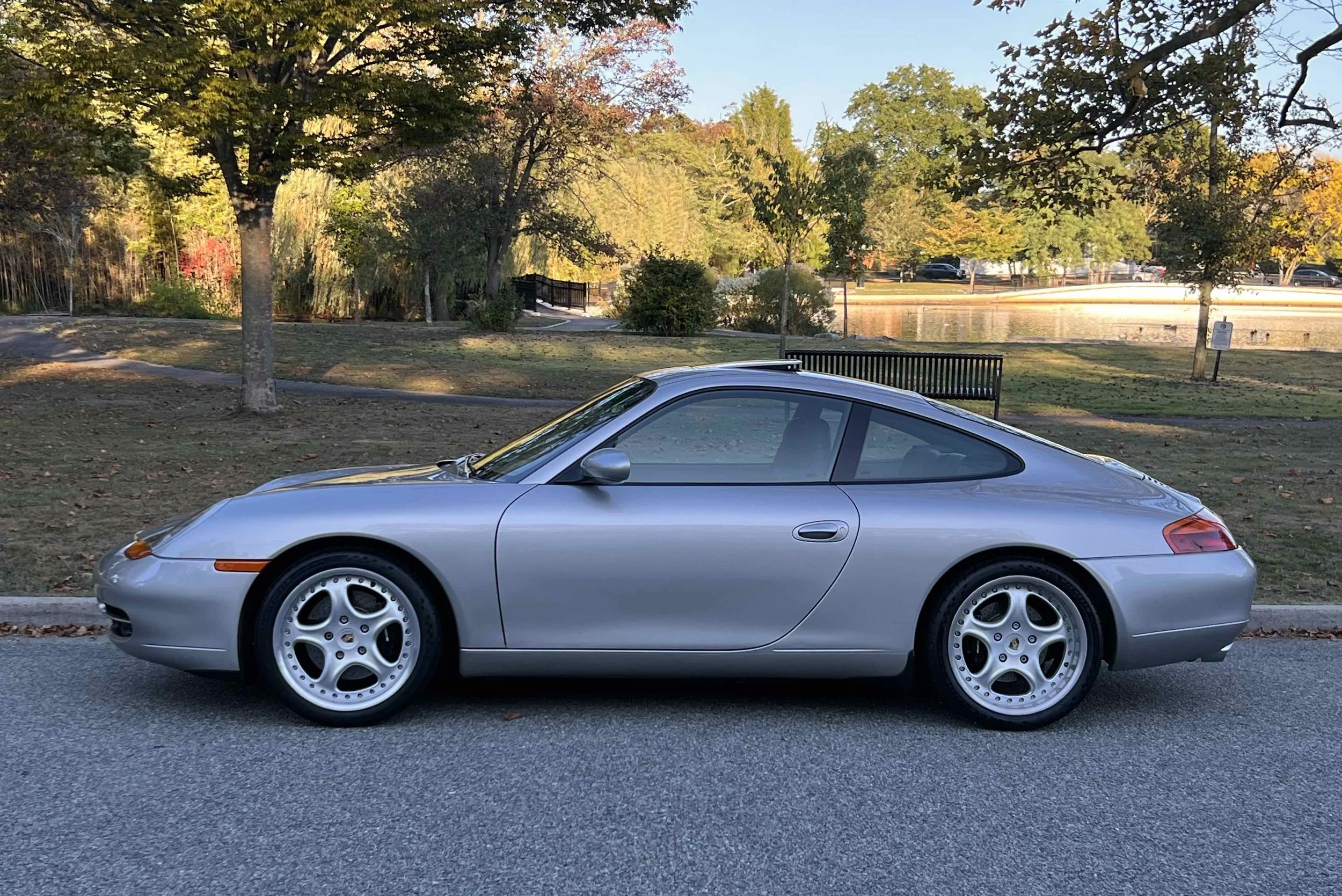 Silver Porsche 911 parked beside a park with trees, a bench, a small lake, and a sign, during autumn