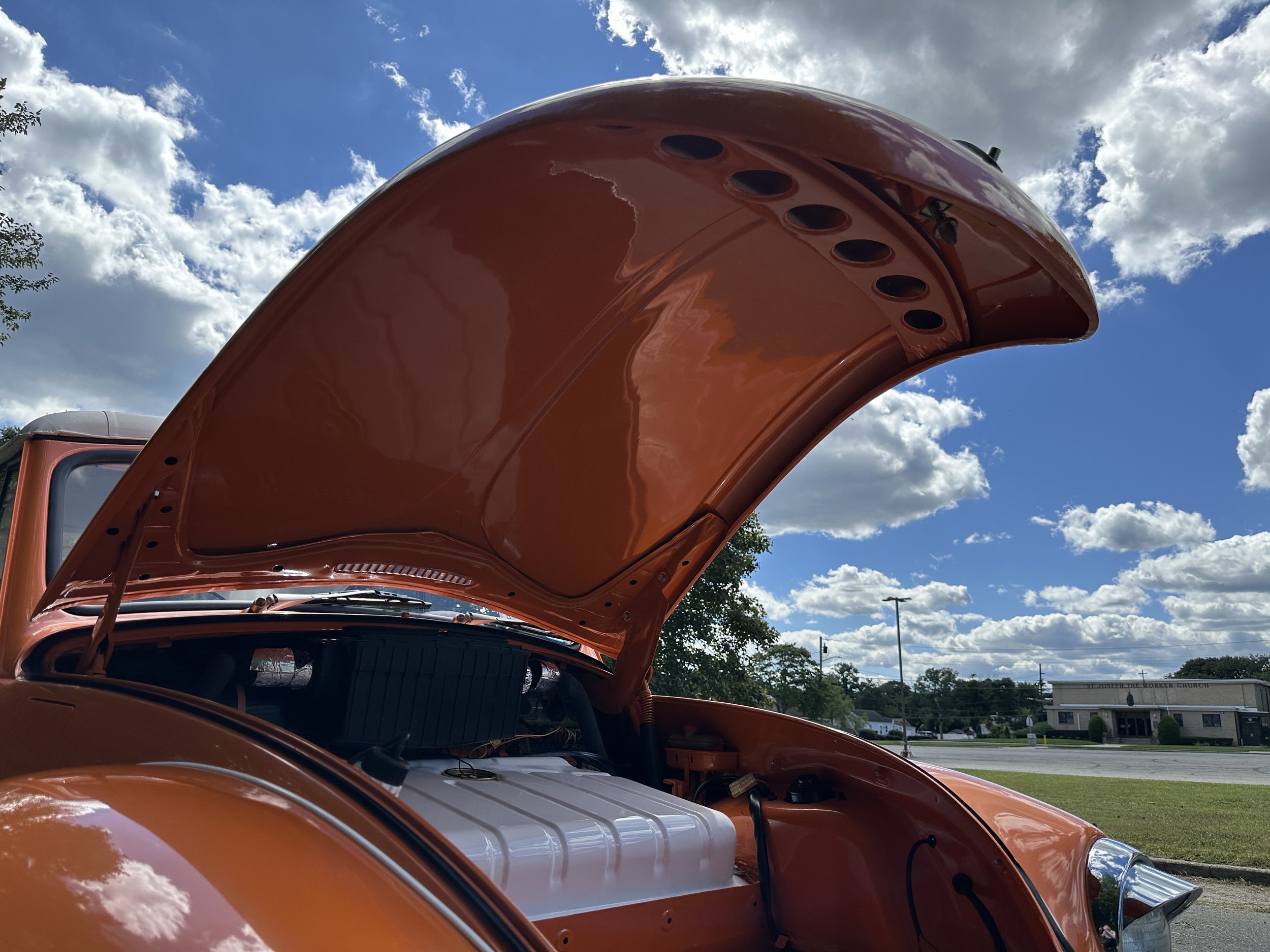 Orange vintage car with open hood showing engine, parked outdoors on a bright sunny day with blue sky and clouds.