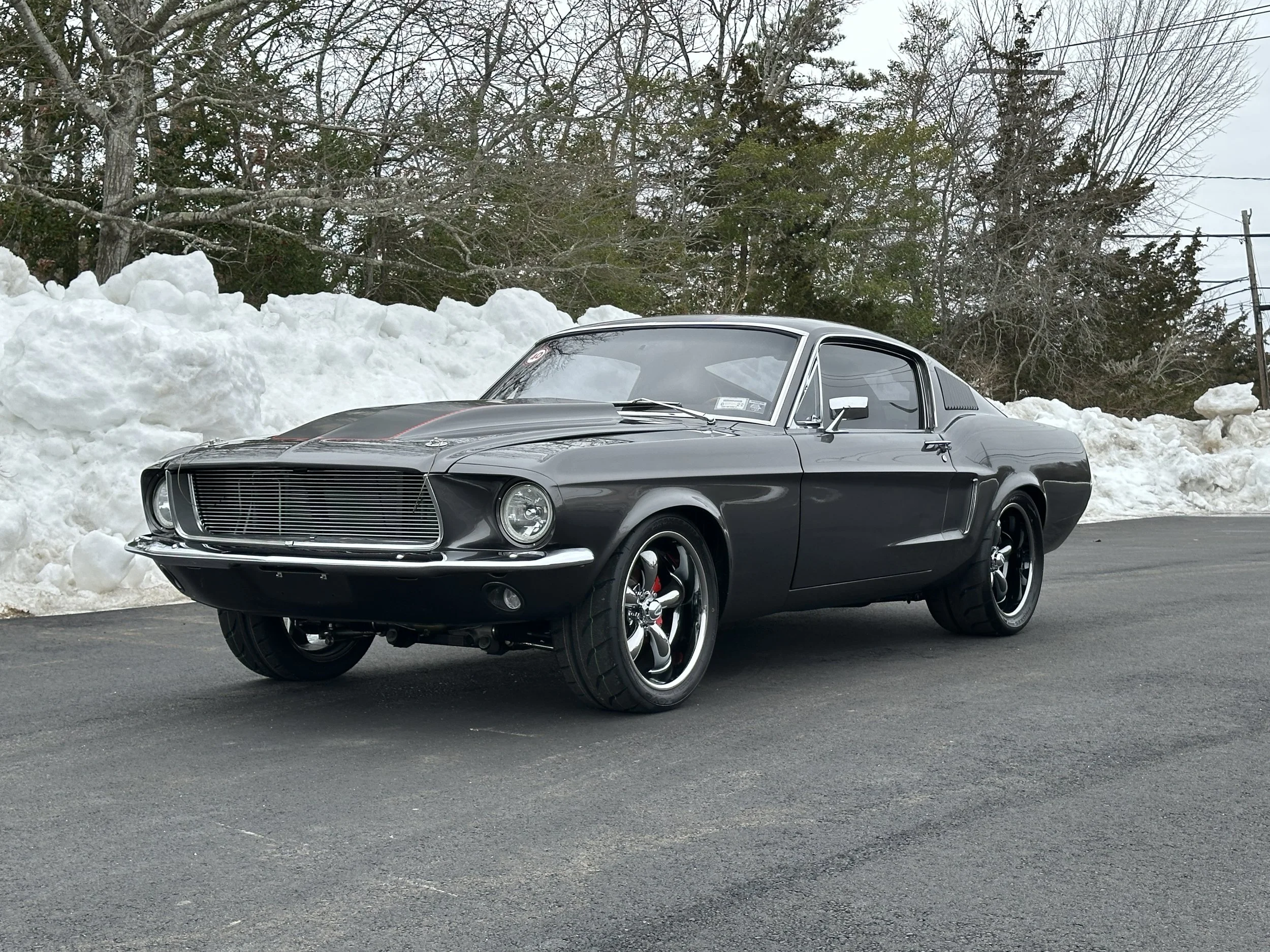 A vintage black muscle car parked on an asphalt surface with snow piled up behind it and leafless trees in the background.