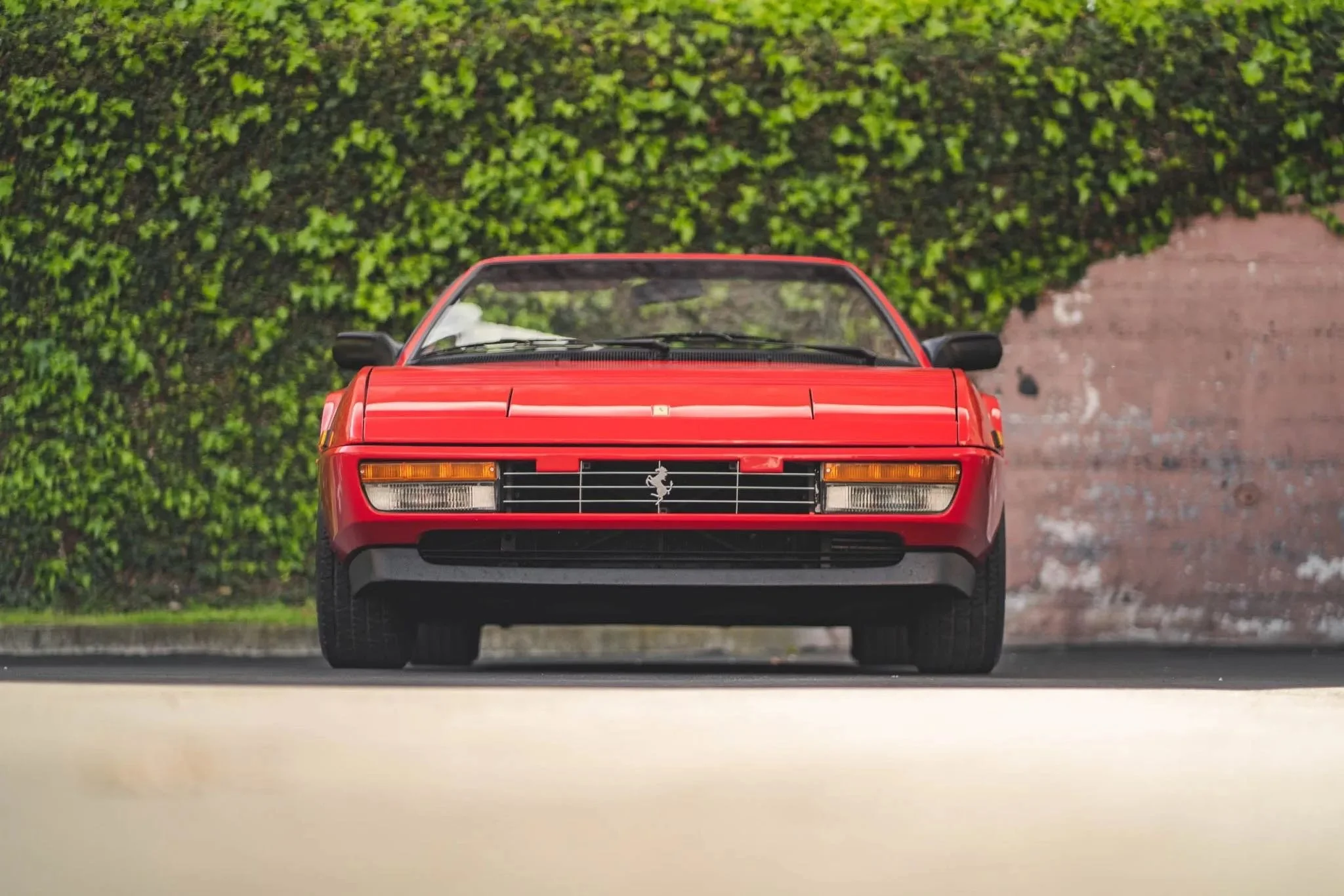 Front view of a vintage red Ferrari sports car parked on a paved surface, with a green leafy wall and a brick wall in the background.