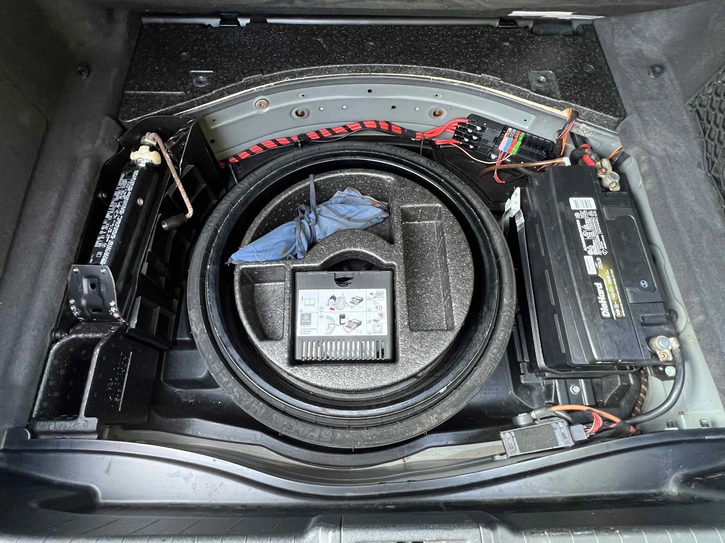 View of a car's trunk, showing a car battery on the right, wiring, and tools. The spare tire well is empty with a blue cloth draped over a small part of the rim.