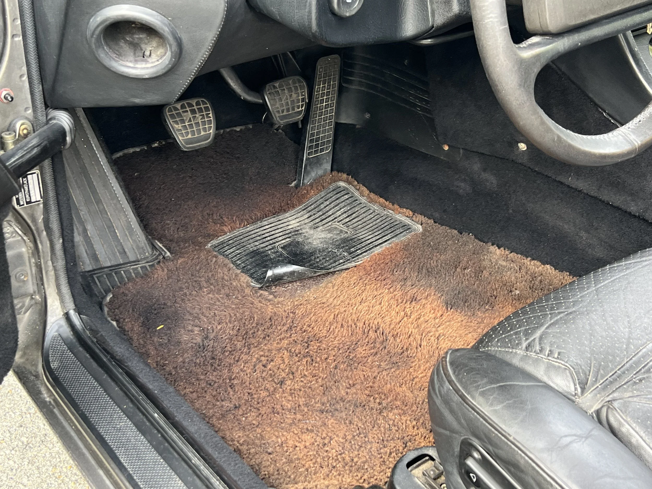 Interior of a car showing a brown and black worn carpeted floor mat, three pedals including clutch, brake, and accelerator, part of the steering wheel, and a black leather seat.